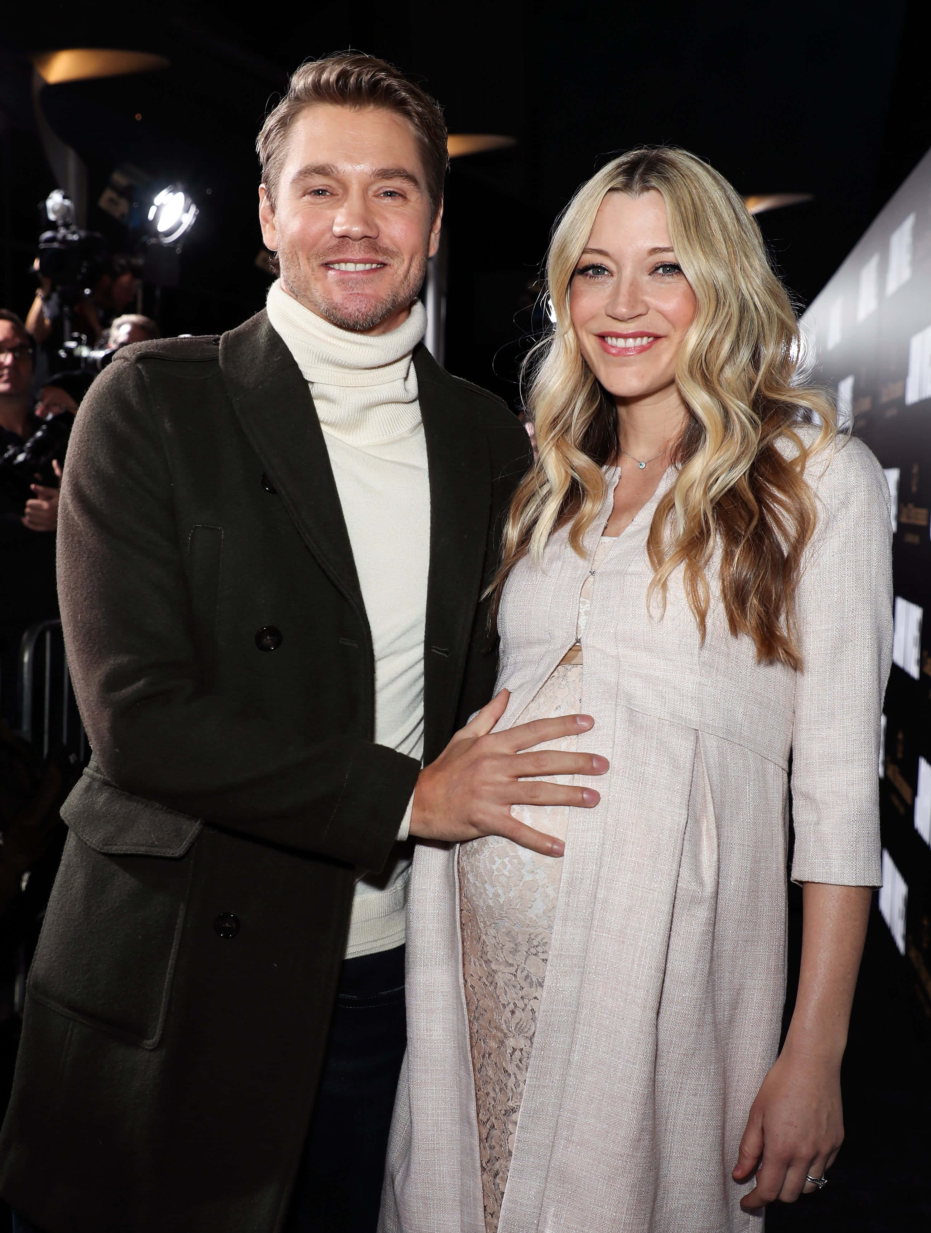 Sarah Roemer 桌布 壁紙 — 一位微笑的孕婦身穿米色連衣裙，站在身穿深色外套的伴侶身旁，身處一個燈光璀璨、相機閃爍的華麗活動中。