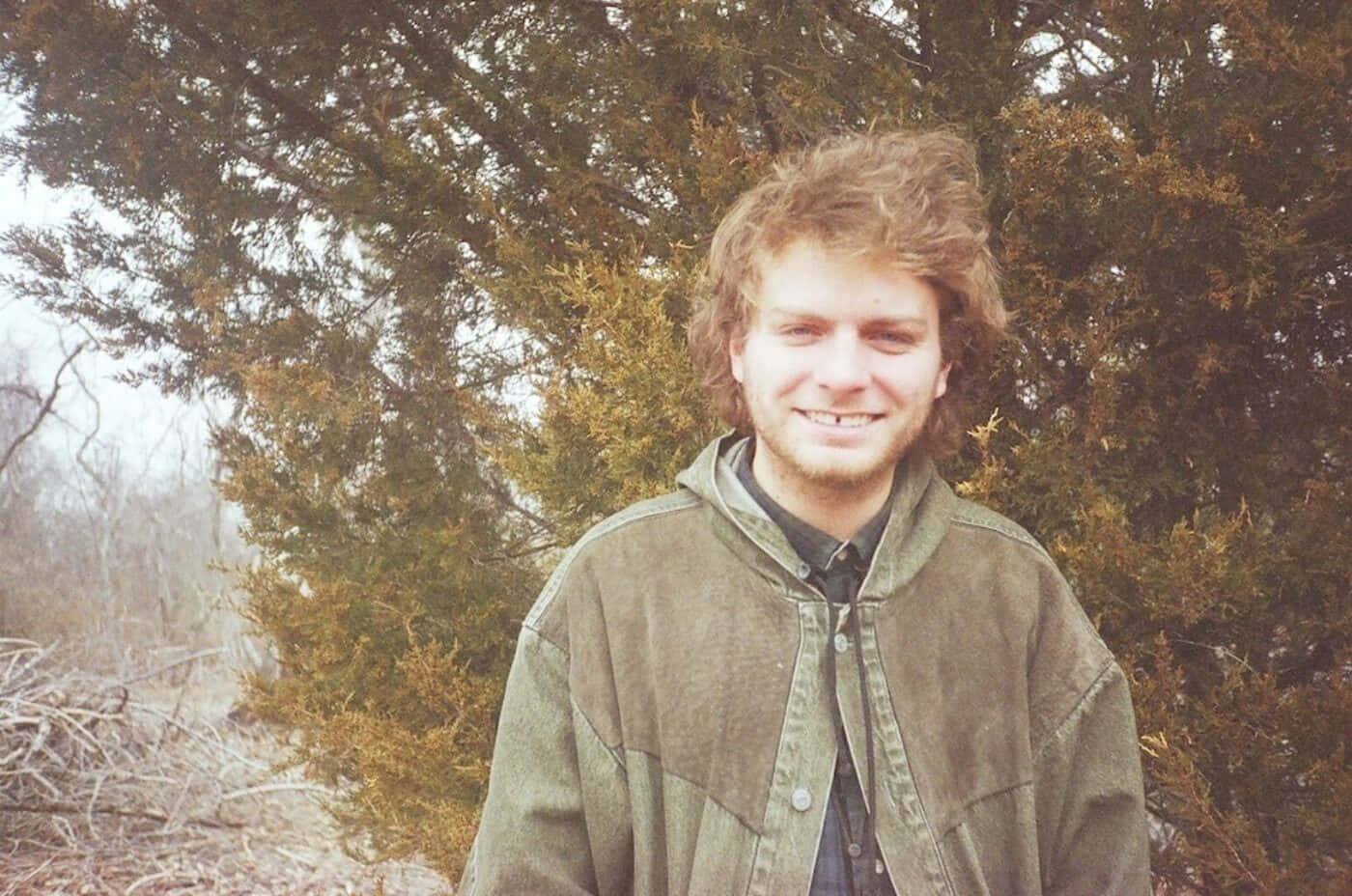  壁紙 — 一個微笑的年輕捲髮男子在涼爽的日子裡，站在戶外茂密的常青樹前。