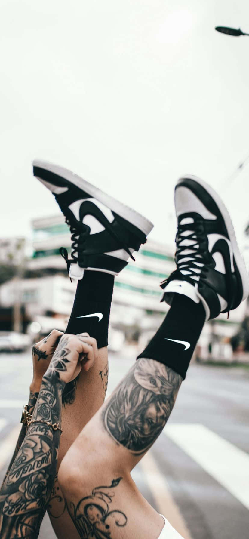  壁紙 — 紋身的雙腿穿著黑色Nike襪子和運動鞋，在模糊的城市街道背景下踢起。