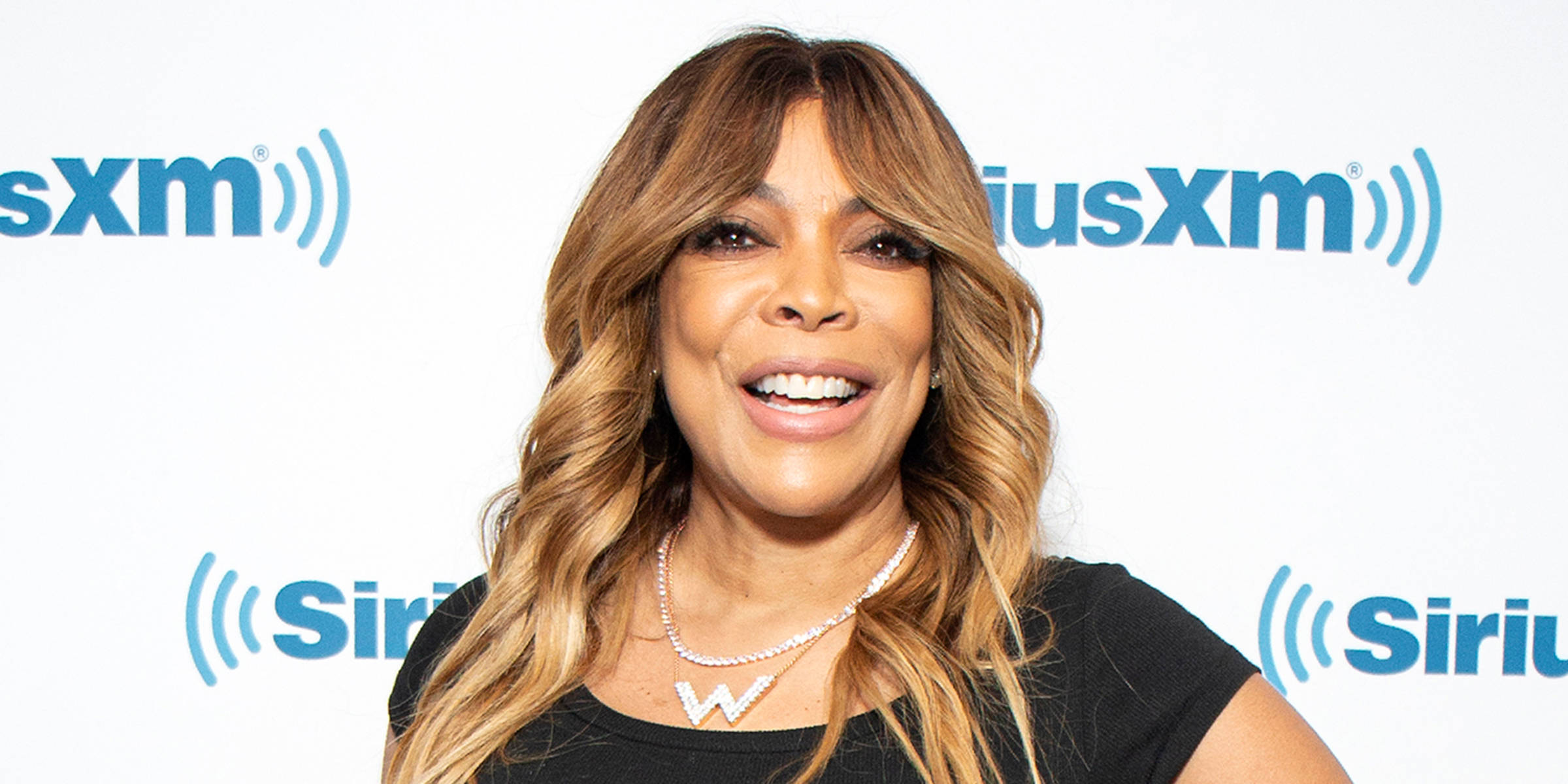 Celebrity 桌布 壁紙 — Wendy Williams 穿著黑色上衣和閃亮的W項鍊，在SiriusXM品牌背景前對著鏡頭燦爛微笑。