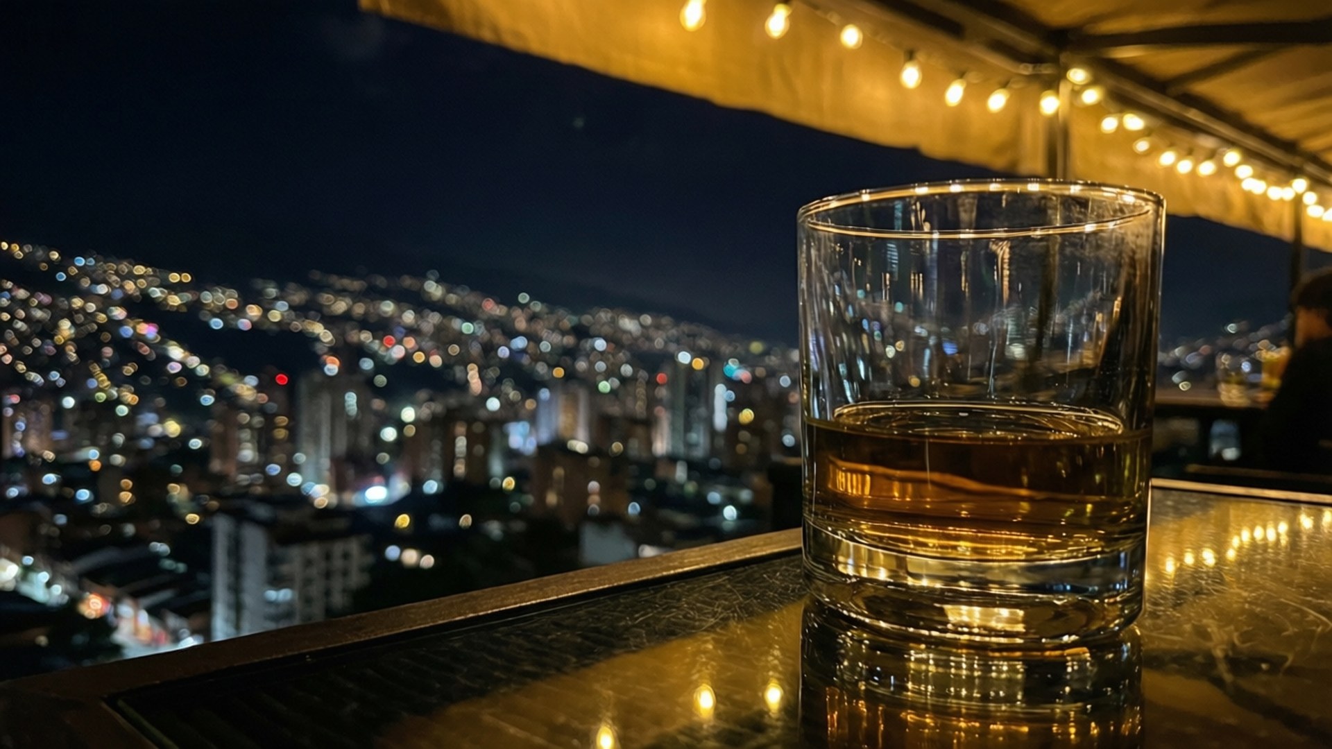 Rooftop Bar Medellín Night 壁紙 — 一杯琥珀色威士忌放在屋頂酒吧的欄杆上，俯瞰夜晚閃爍的城市景觀。