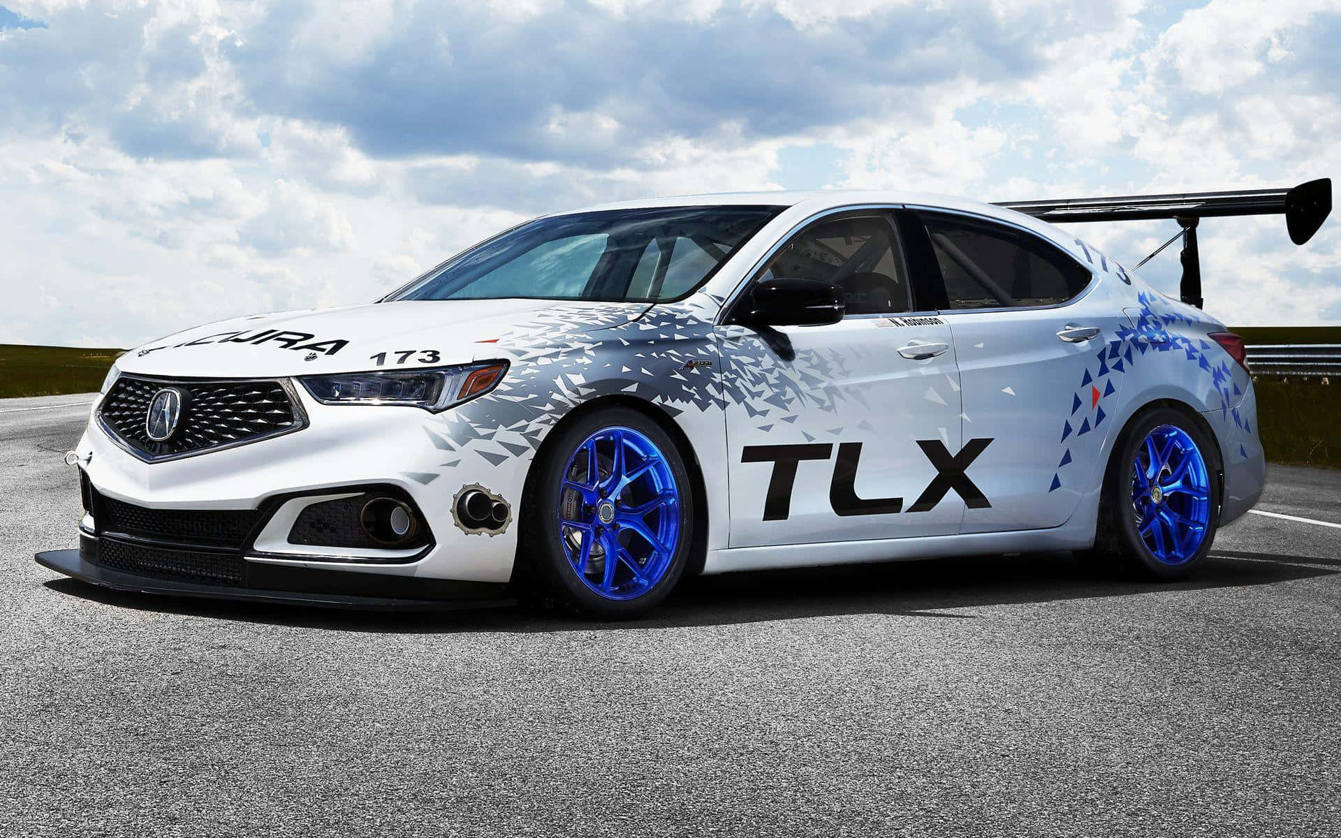 Acura Tlx 桌布 壁紙 — 一輛白色Acura TLX賽車，配備藍色輪圈和幾何圖案貼紙，在多雲的天空下於賽道上奔馳。