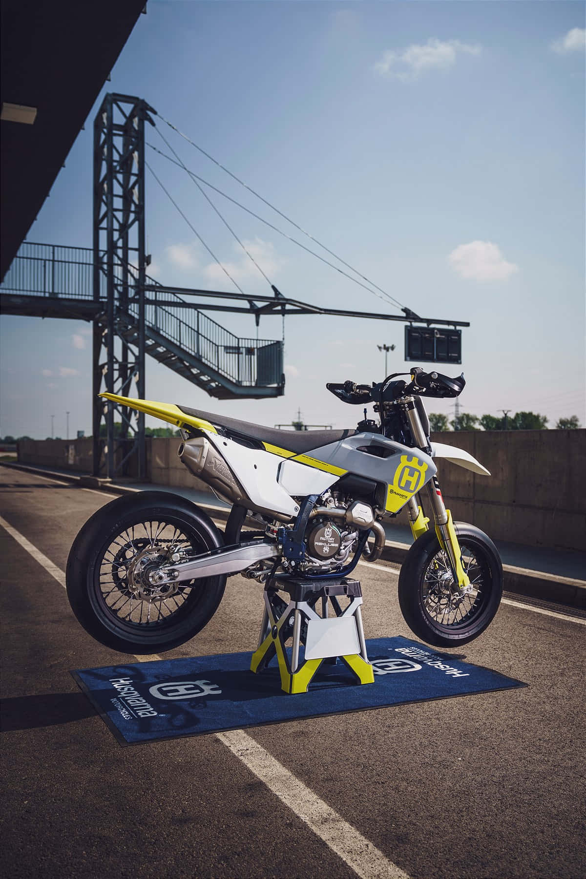  壁紙 — 一輛白色和黃色的 Husqvarna 越野摩托車，在賽車場的藍色品牌墊上，於晴朗的藍天下展示。
