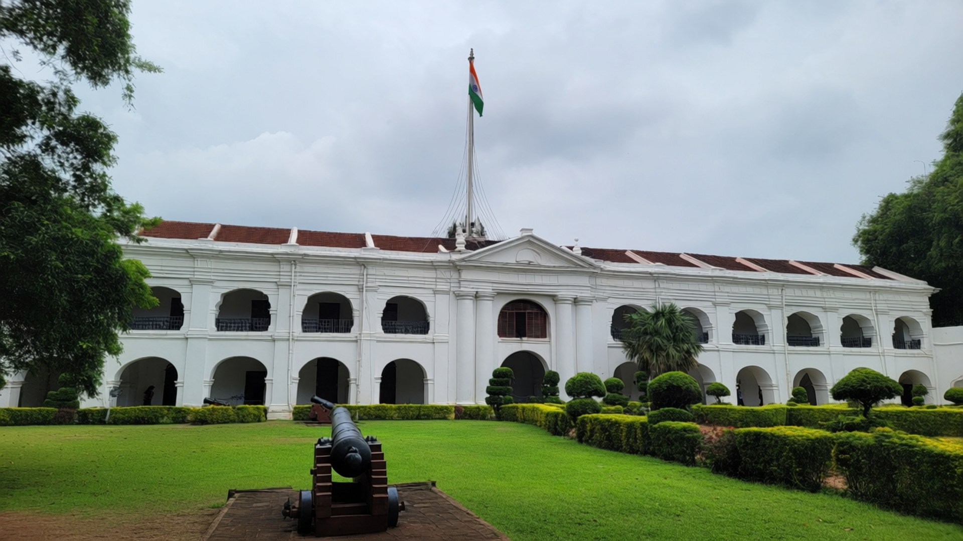 Fort St. George Pride 壁紙 — 一座宏偉的白色殖民風格建築，帶有拱形窗戶和懸掛印度國旗的旗杆，矗立在修剪整齊的草坪後面，前景有一門大砲。