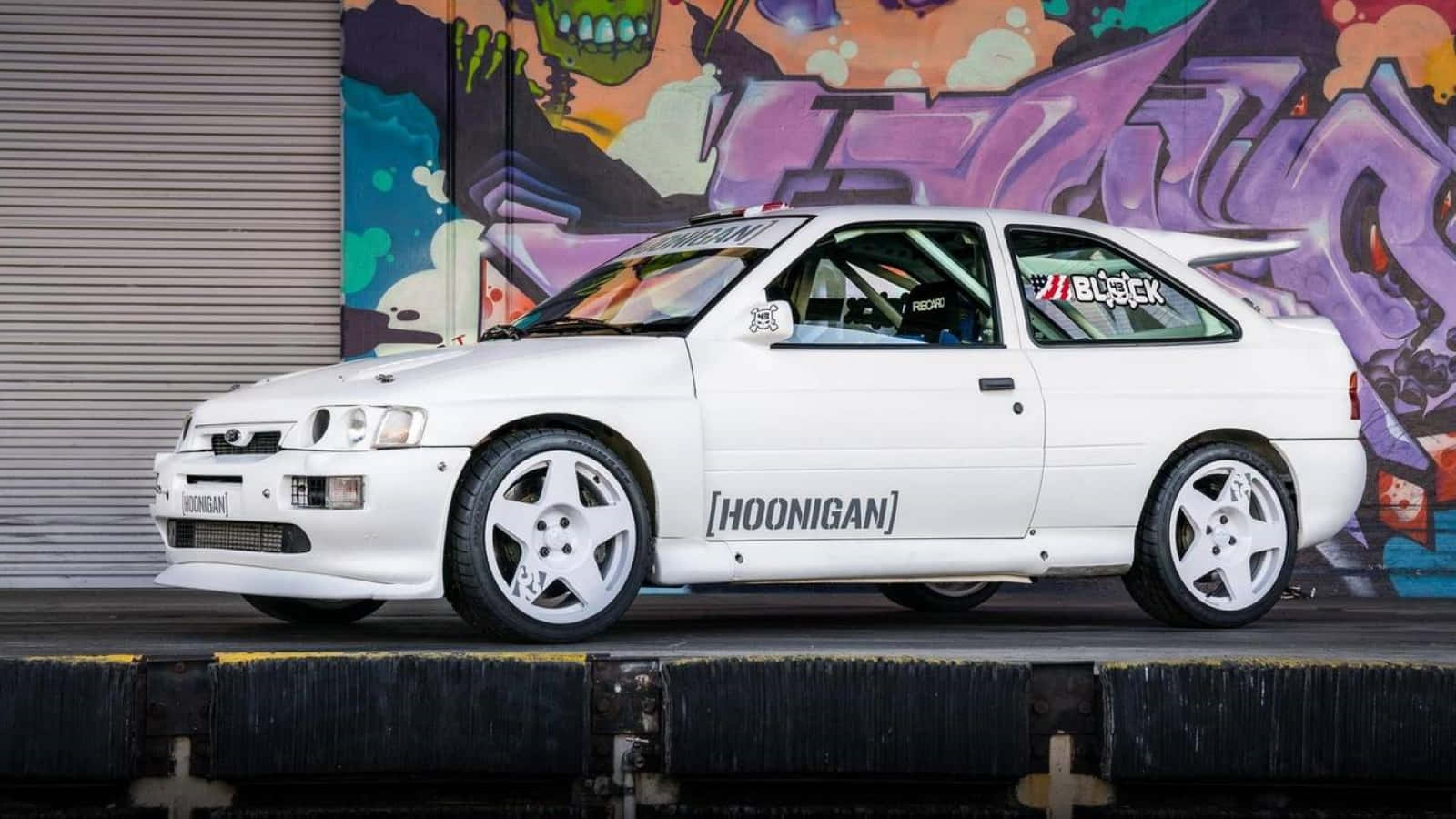 Ford Escort 桌布 壁紙 — 一輛白色Hoonigan拉力賽車停放在充滿活力的塗鴉牆前，牆上有大膽的紫色和橙色標籤。