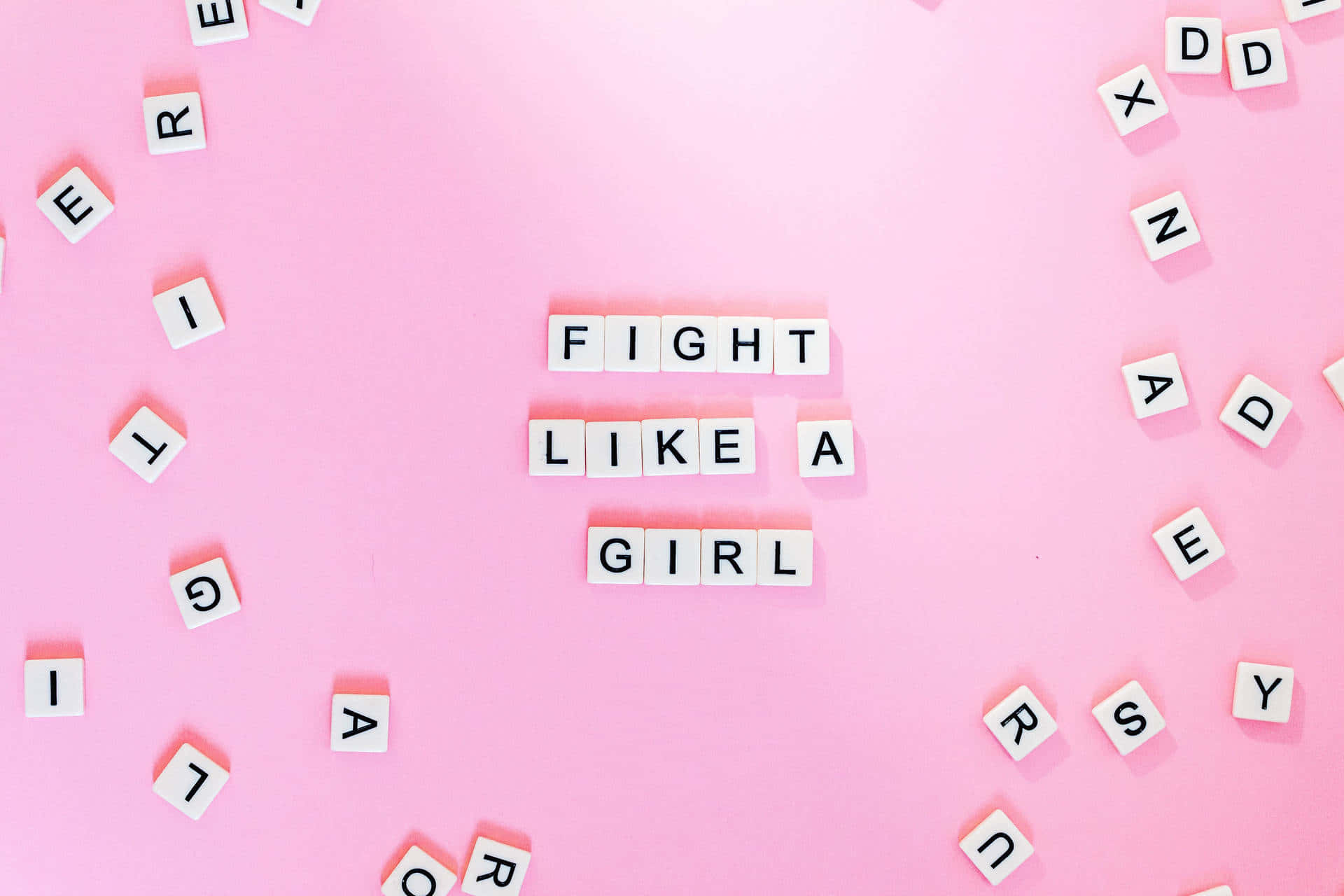  壁紙 — 白色字母瓷磚在柔和的粉紅色背景中央拼出「FIGHT LIKE A GIRL」，周圍散落著字母。
