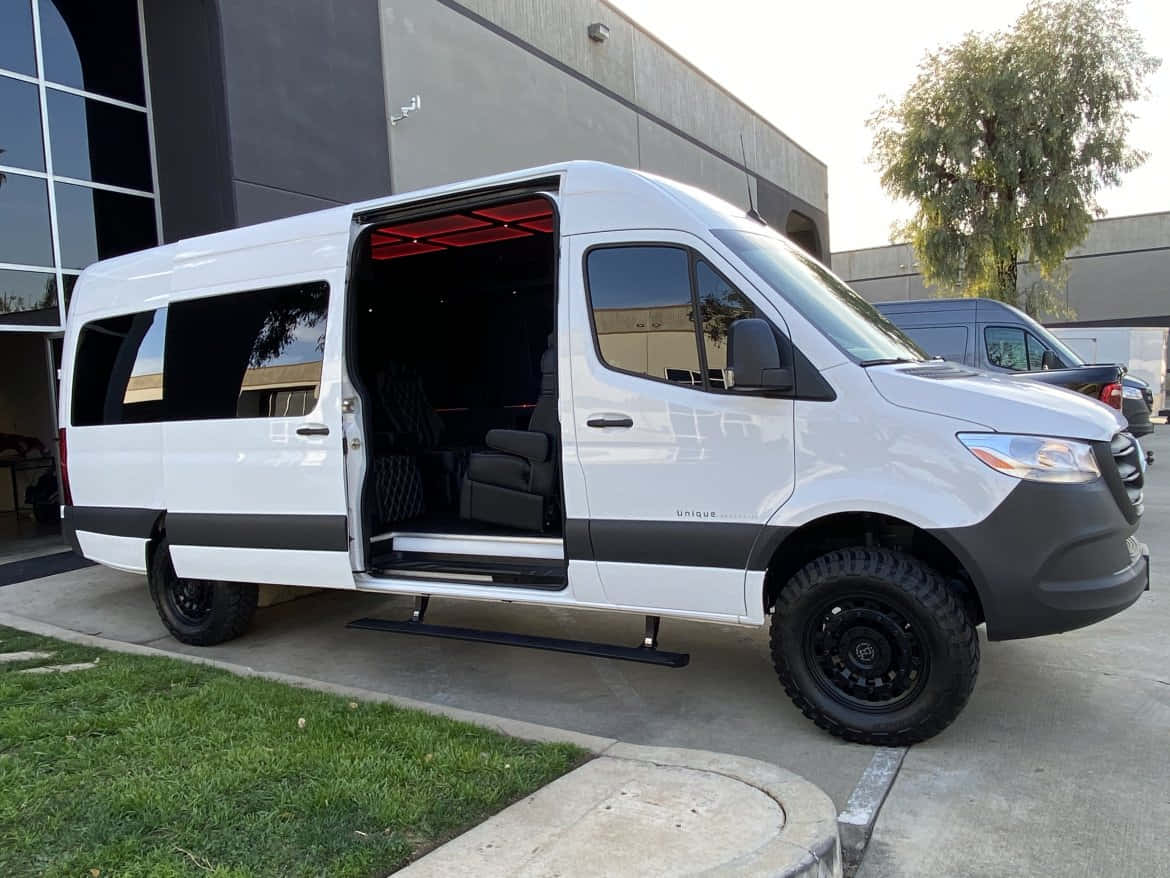 Mercedes Benz Sprinter 桌布 壁紙 — 一輛白色豪華廂型車，帶有黑色飾邊和堅固輪胎，停在一棟現代建築外，側門打開，露出紅色內部照明。