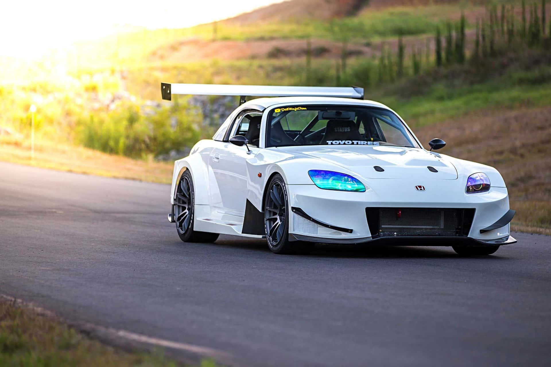  壁紙 — 一輛白色改裝本田S2000賽車，帶有發光的藍色車頭燈，在黃金時段沿著蜿蜒的鄉村道路飛馳。