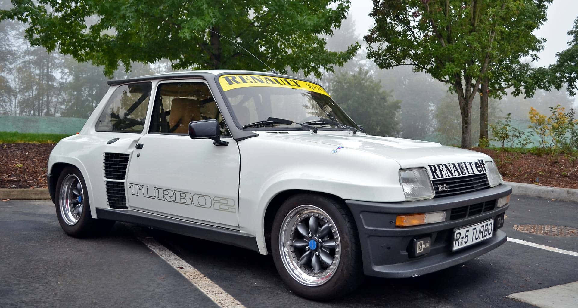 Renault 5 Turbo 桌布 壁紙 — 一輛白色雷諾5 Turbo 2拉力賽車停放在霧氣瀰漫、樹木成排的停車場，背景同樣霧濛濛。
