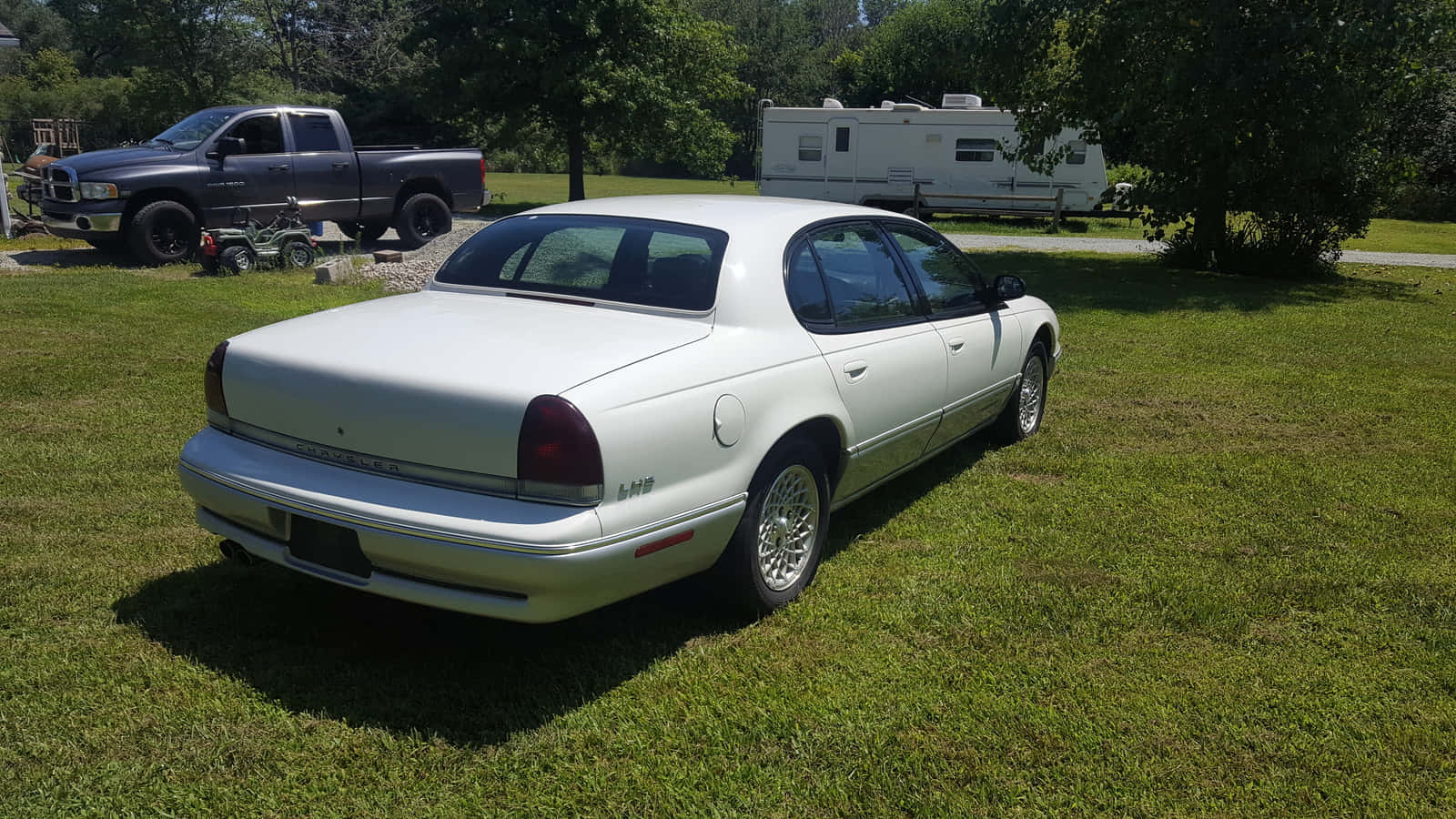 Chrysler Lhs 桌布 壁紙 — 一輛白色轎車停在草地上，背景有一輛黑色皮卡車和露營車，在明亮的陽光下。