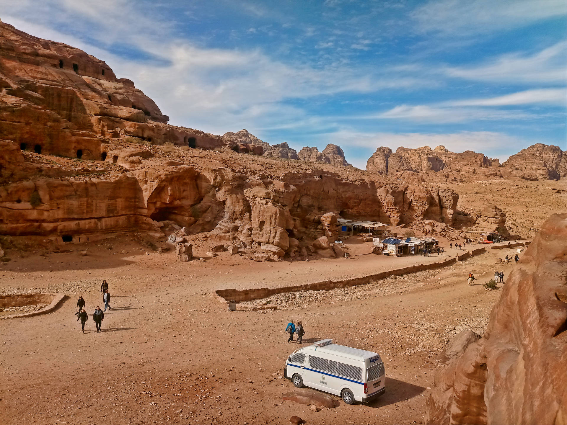 白色廂型車停放在沙漠峽谷中，有遊客和古老的岩石住所 — Jordan 壁紙
