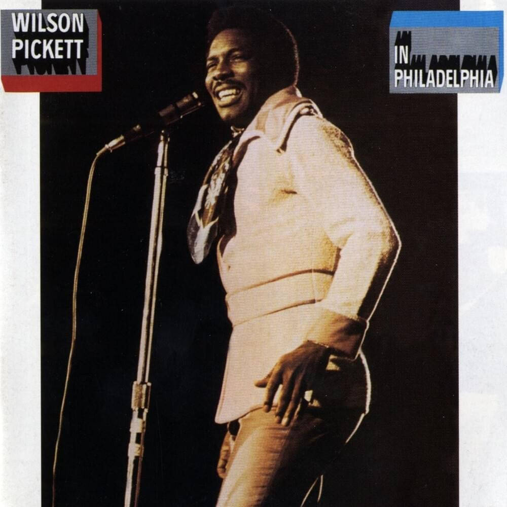 音樂 桌布 壁紙 — Wilson Pickett 在燈光昏暗的舞台上對著麥克風唱歌，露出愉快的笑容。