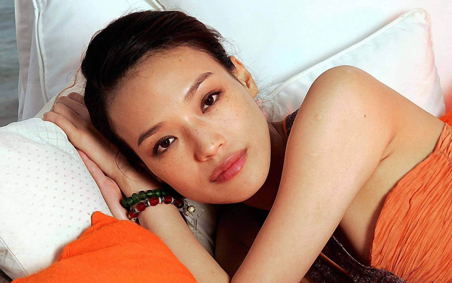Shu Qi 桌布 壁紙 — 一位深色頭髮的女性將頭靠在白色枕頭上，穿著橙色上衣和珠子手鐲。