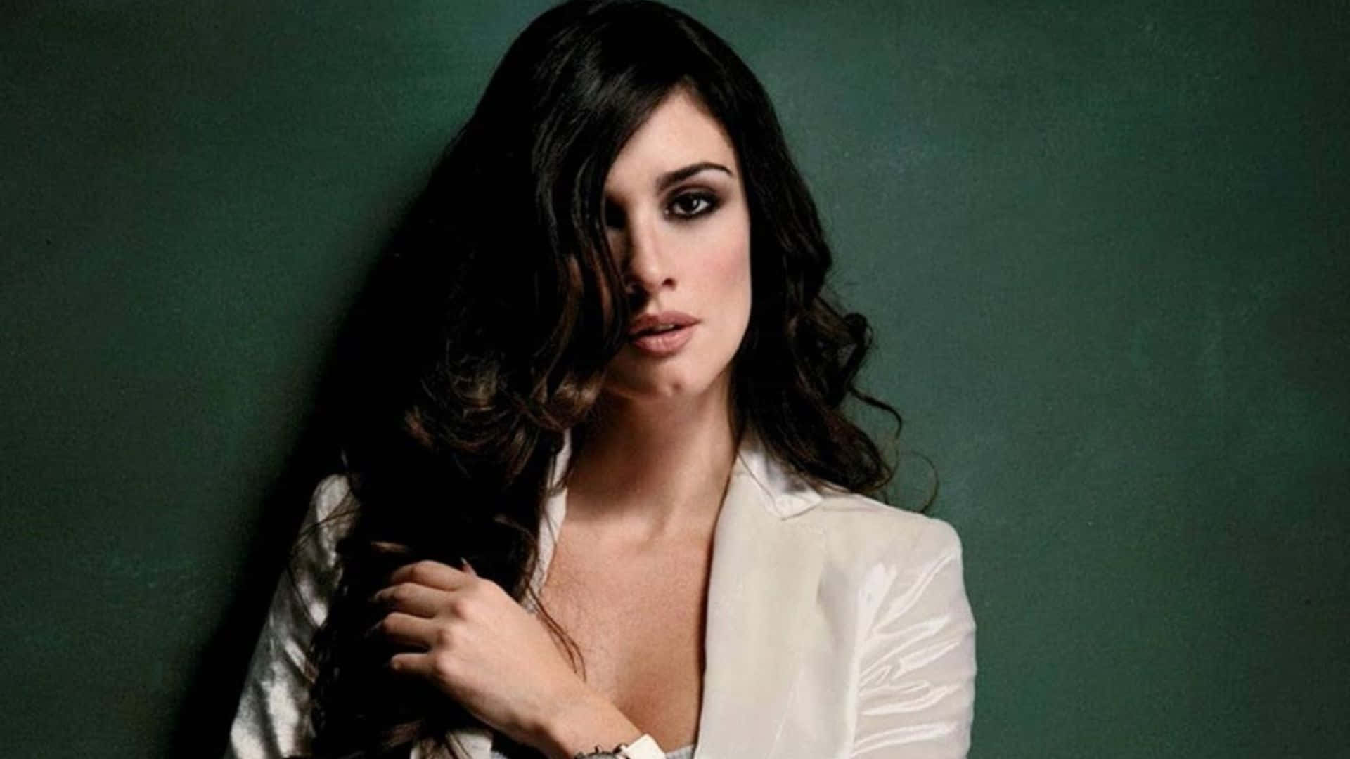 Paz Vega 桌布 壁紙 — 一位擁有深色波浪髮和煙燻妝眼睛的女性，在柔和的綠色背景下，觸摸頭髮並投以強烈的凝視。