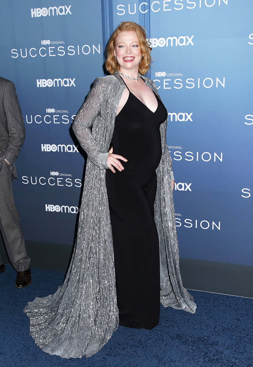 Sarah Snook 桌布 壁紙 — 一位身穿黑色連衣裙和閃亮銀色披肩的女性在Succession活動的藍色地毯上自信地擺姿勢。