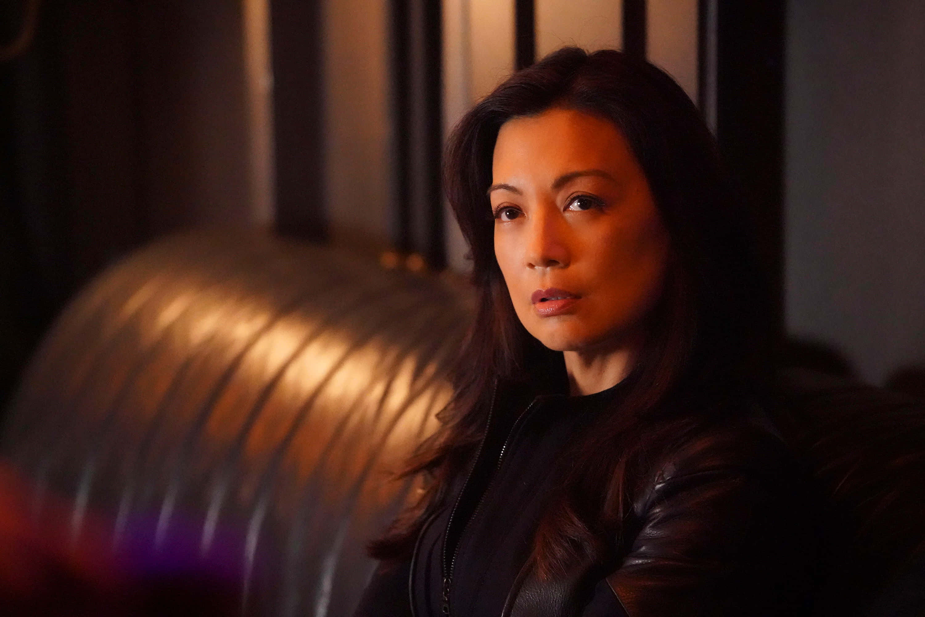 Ming Na Wen 桌布 壁紙 — 一位身穿黑色皮夾克的女性，在昏暗的暖光中坐著，表情嚴肅。