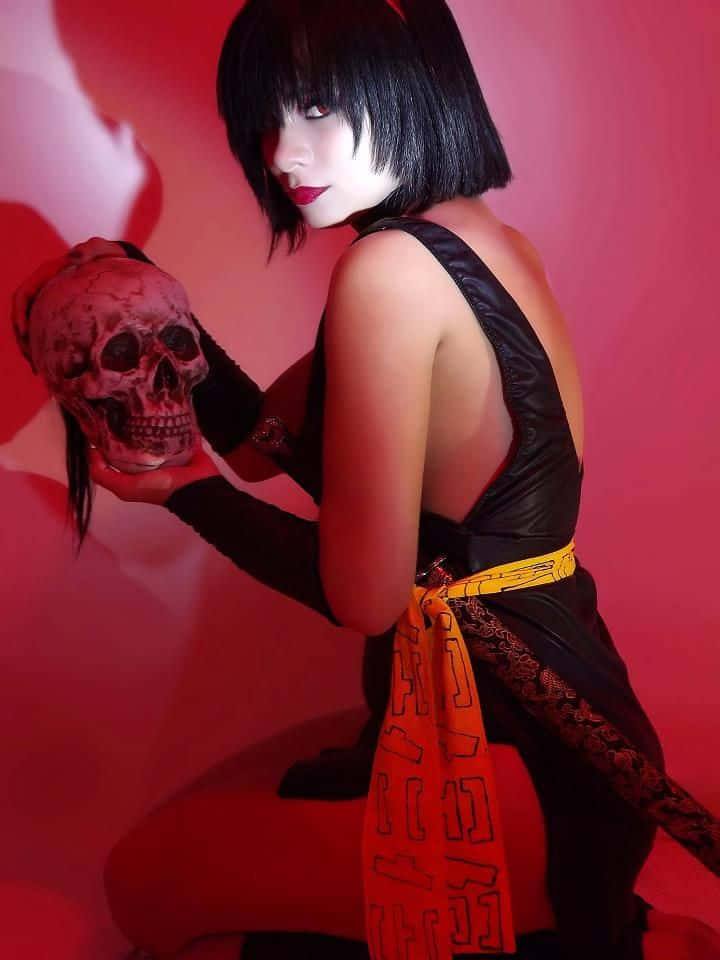 Inuyasha Yura 桌布 壁紙 — 一位留黑色鮑伯頭、紅唇的女性，在鮮紅色背景前手持頭骨，身穿黑色服裝，搭配大膽的橙色圖案腰帶。