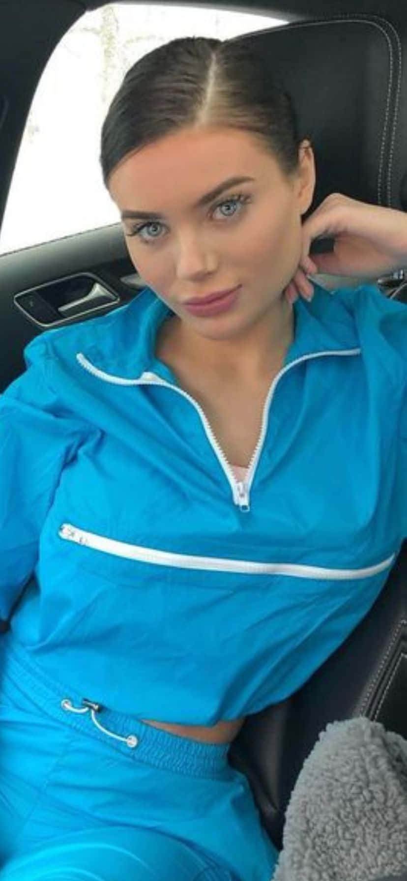  壁紙 — 一位身穿亮藍色運動服的女性自信地坐在汽車座椅上，一隻手靠近脖子，直視鏡頭。