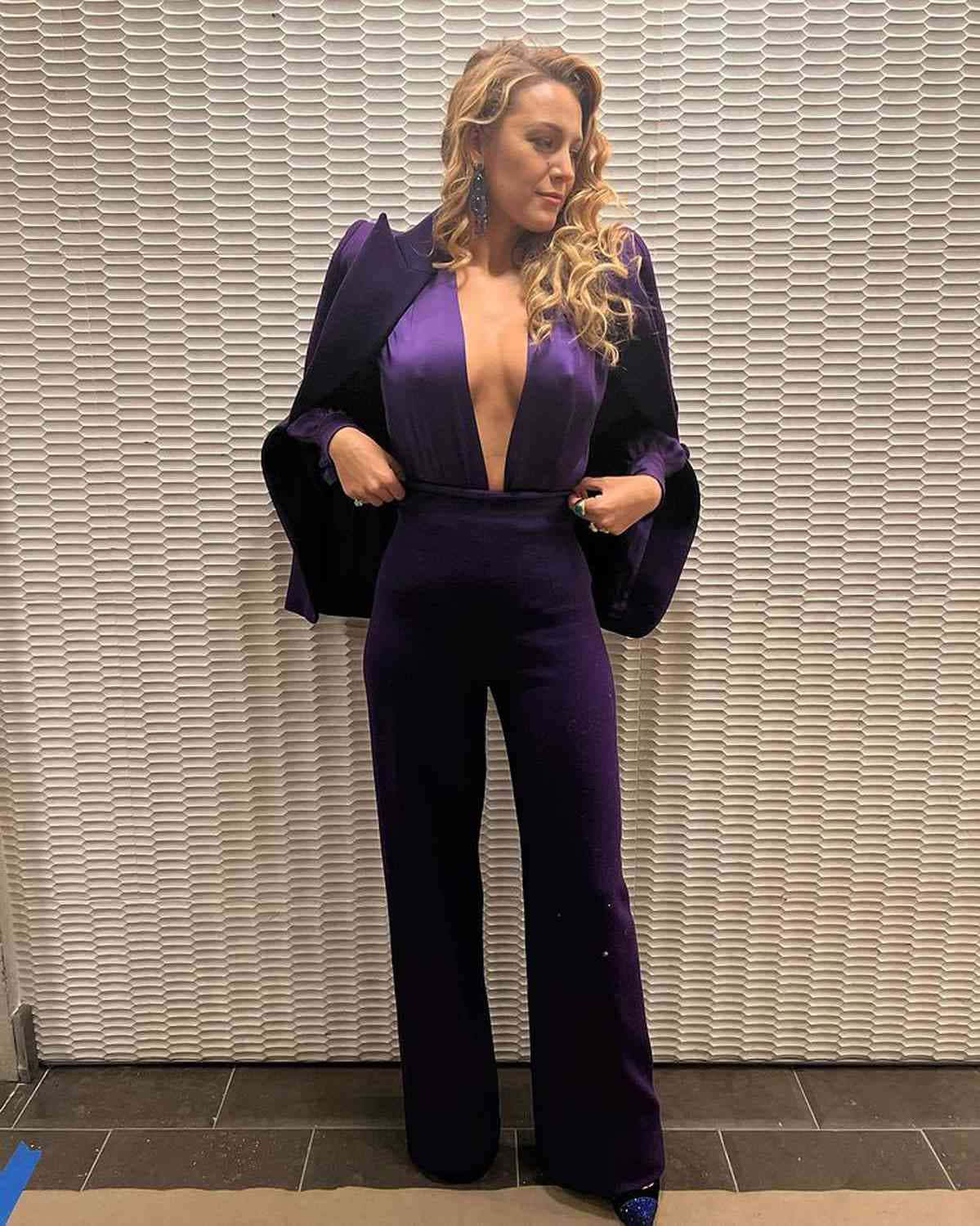 Purple Suit 桌布 壁紙 — 一位身穿深紫色天鵝絨連身褲、領口低垂的女性，自信地站在有紋理的白色牆壁前。