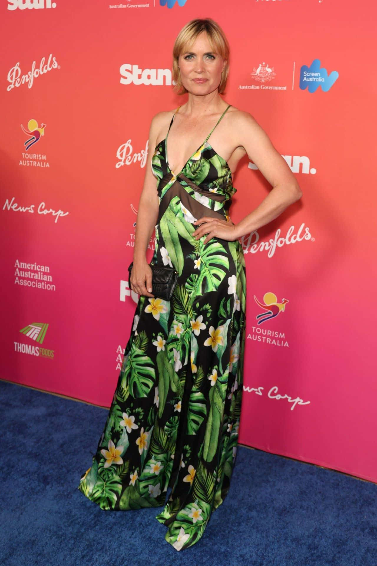 Radha Mitchell 桌布 壁紙 — 一位穿著熱帶印花長裙的女性在藍色地毯上自信地擺姿勢，背景是充滿活力的粉紅色品牌背景。