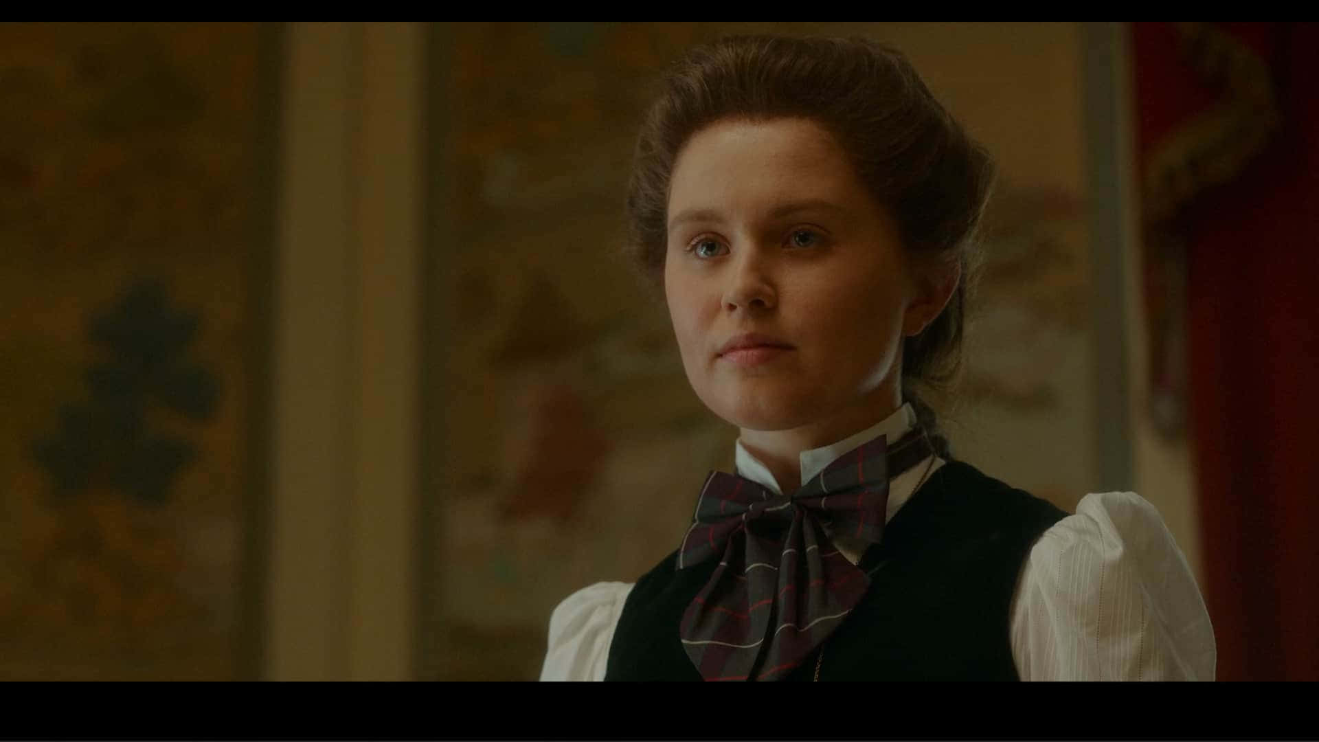 Eliza Scanlen 桌布 壁紙 — 一位穿著復古服裝、戴著格子蝴蝶結的女性，在溫暖燈光的室內若有所思地凝視。