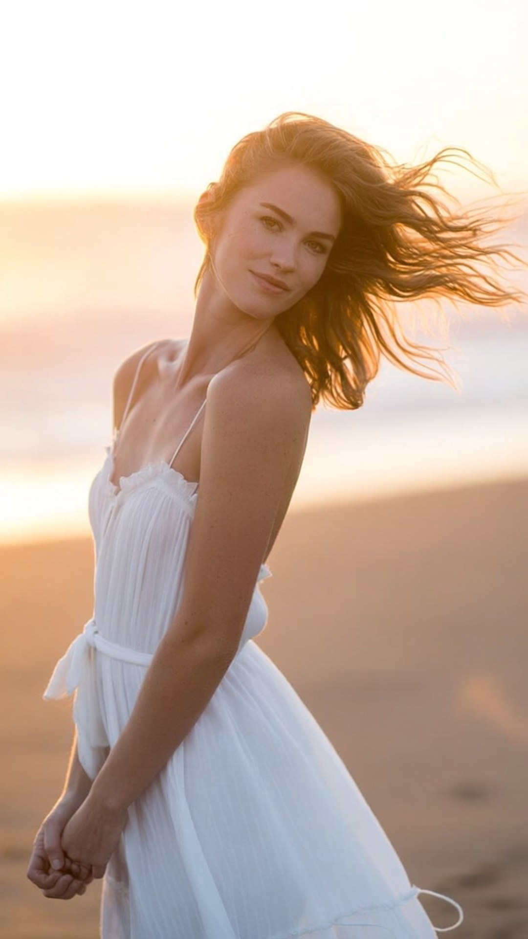 Beach Sunset Serenity 壁紙 — 一位身穿飄逸白色連衣裙的女性在日落時分站在沙灘上，風吹動她的頭髮，她回頭看向肩膀後方。