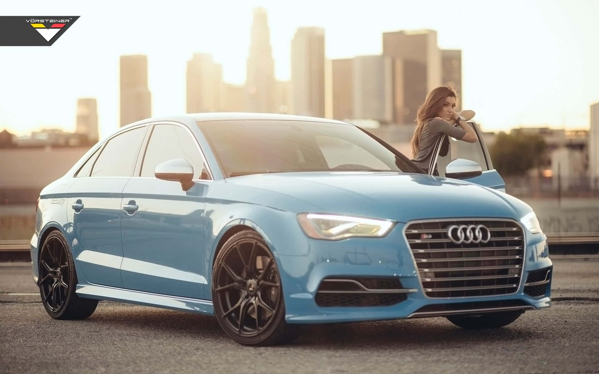 Audi S3 桌布 壁紙 — 一名女士從一輛停在城市街道上的淺藍色奧迪轎車中探出身子，背景是金色時刻朦朧的城市天際線。