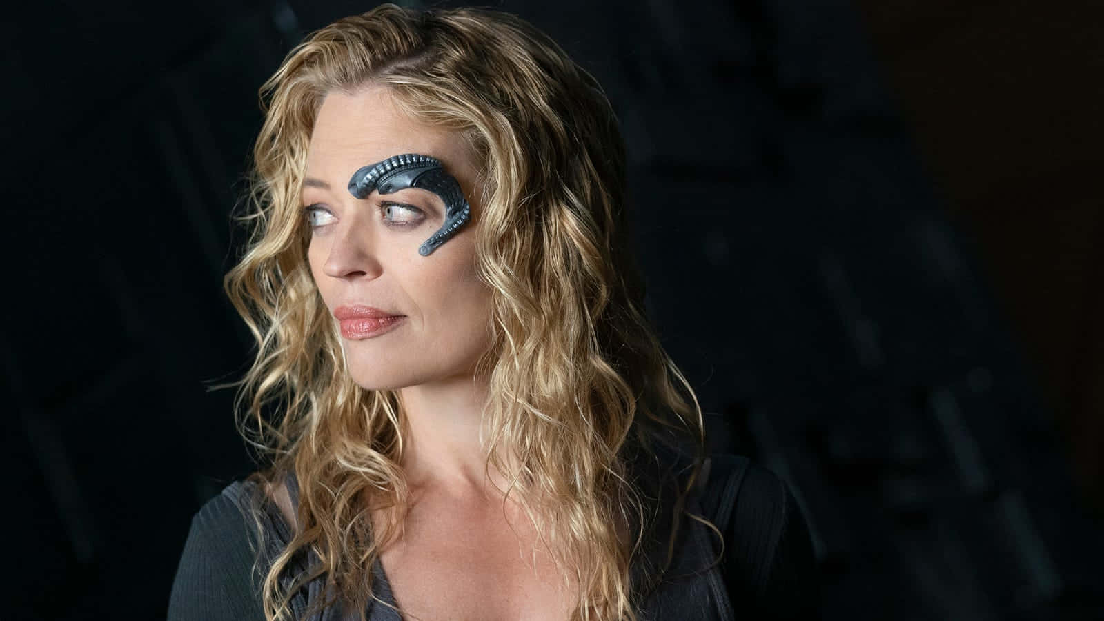 Jeri Ryan 桌布 壁紙 — 一位擁有波浪形金髮和生物力學外星人面部植入物的女性，在黑暗背景下若有所思地凝視。