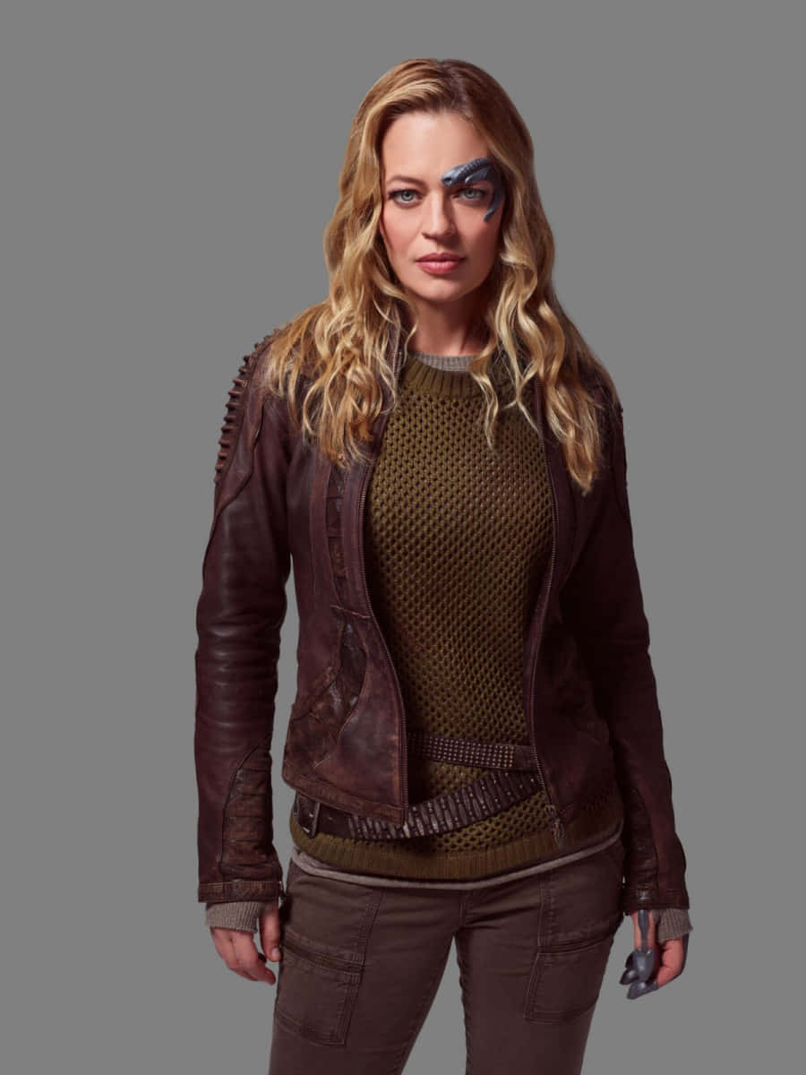 Jeri Ryan 桌布 壁紙 — 一位擁有波浪金髮和藍色臉部彩繪的女性，在灰色背景前穿著棕色皮夾克，內搭有紋理的毛衣。