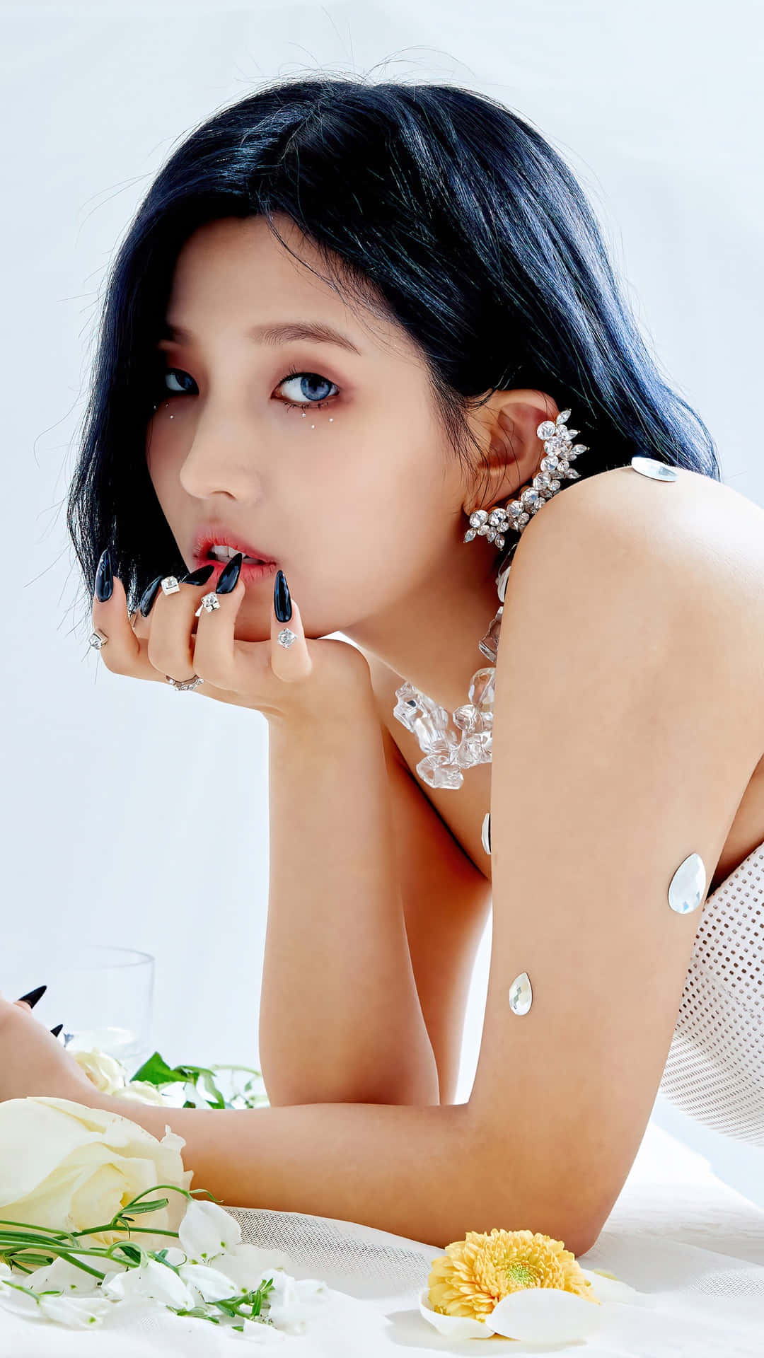  壁紙 — 一位深色頭髮、藍色眼睛的女性，將下巴擱在飾有黑色指甲和戒指的手上，周圍環繞著白色和黃色的花朵，背景是明亮的白色。