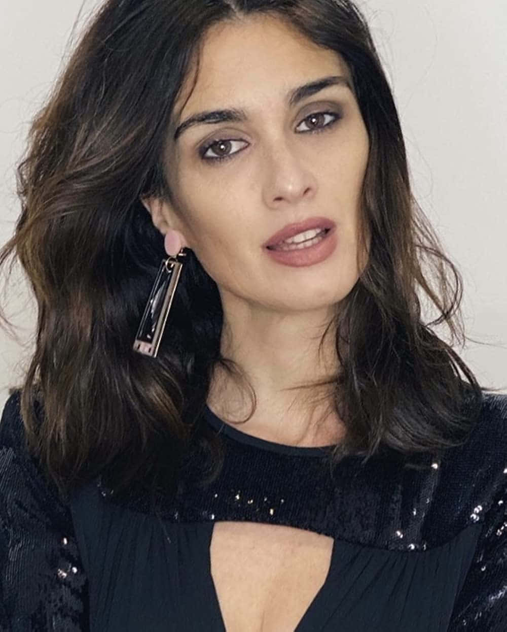 Paz Vega 桌布 壁紙 — 一位擁有深色波浪髮型和誇張妝容的女性，在純白背景前穿著黑色亮片上衣和大型幾何耳環。