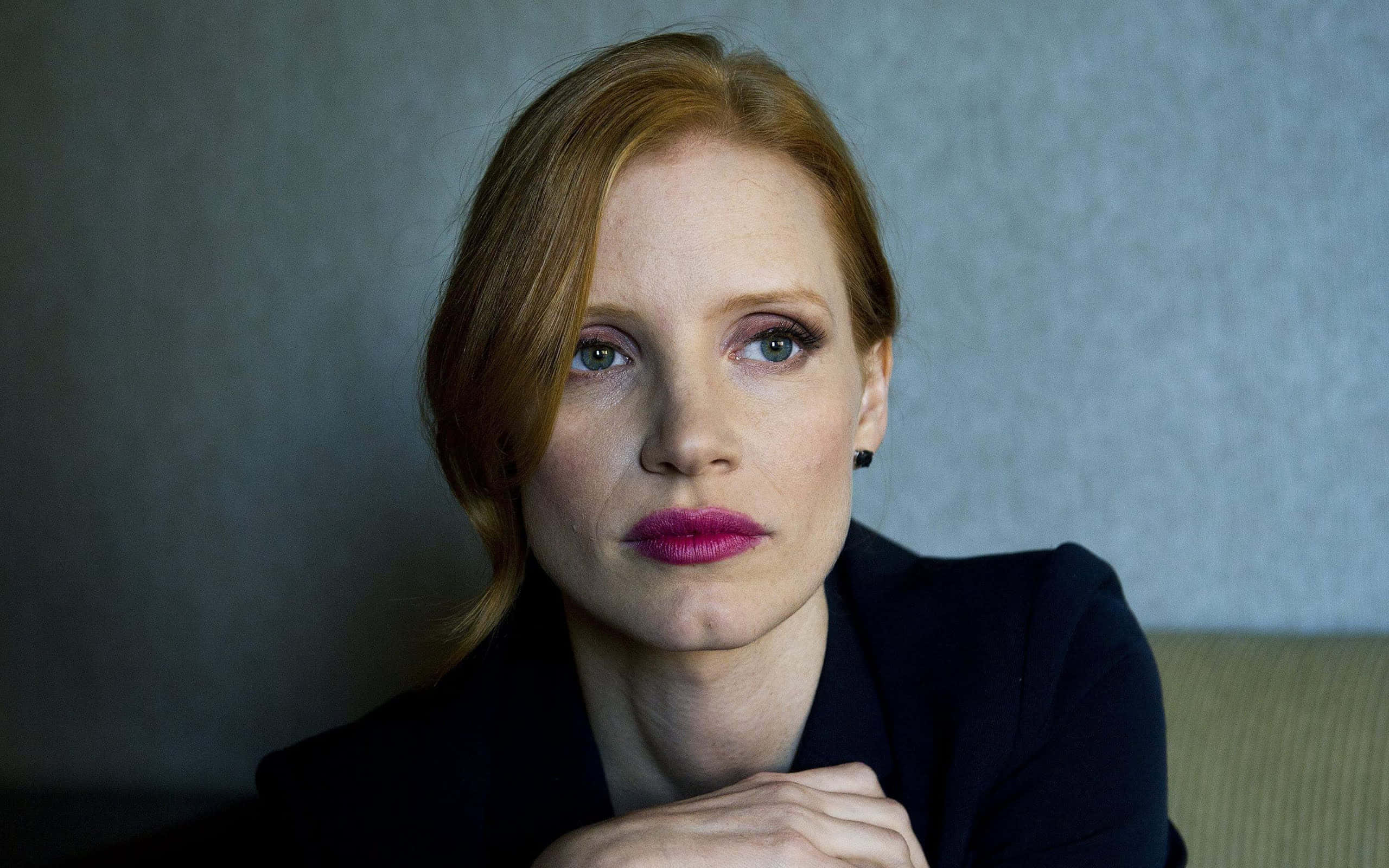 jessica chastain 桌布 壁紙 — 一位紅髮、嘴唇大膽粉紅的女性，專注地凝視著一面柔和灰色紋理牆。