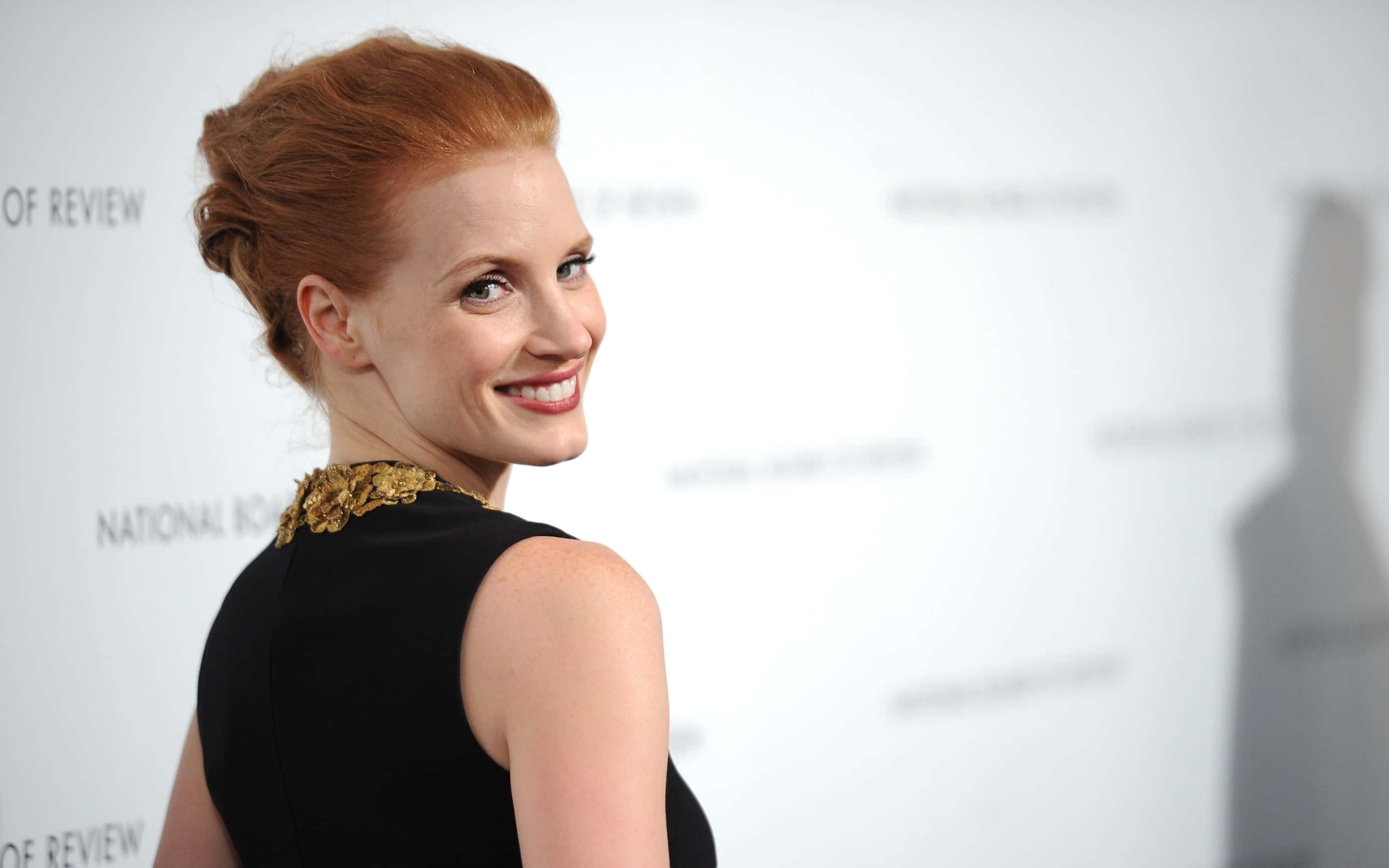 jessica chastain 桌布 壁紙 — 一位紅髮盤起的女士，身穿金色裝飾領口的黑色連衣裙，回頭微笑。