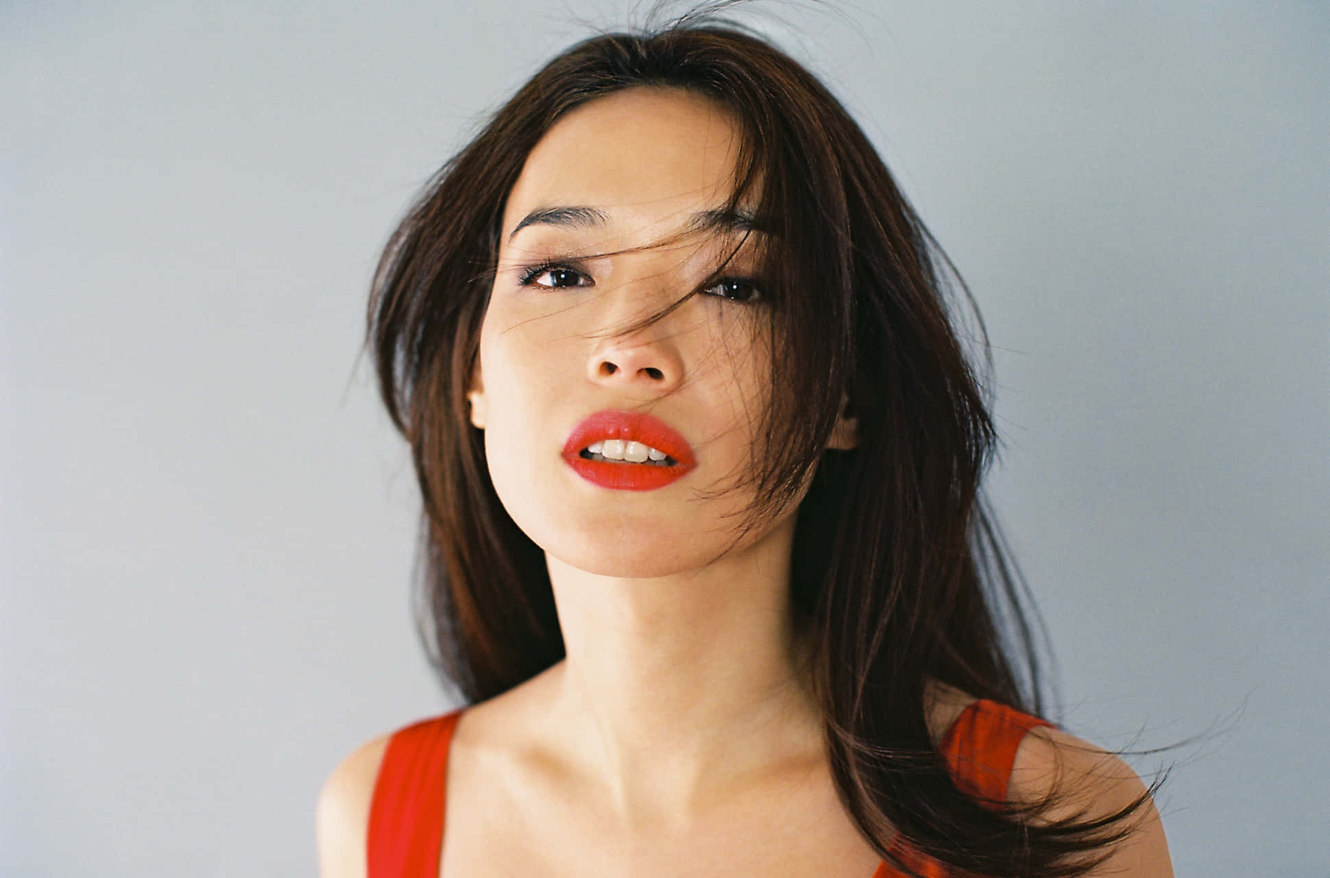 Shu Qi 桌布 壁紙 — 一位擁有鮮豔紅唇和隨風飄揚的深色頭髮的女性，在純灰色背景下強烈凝視。