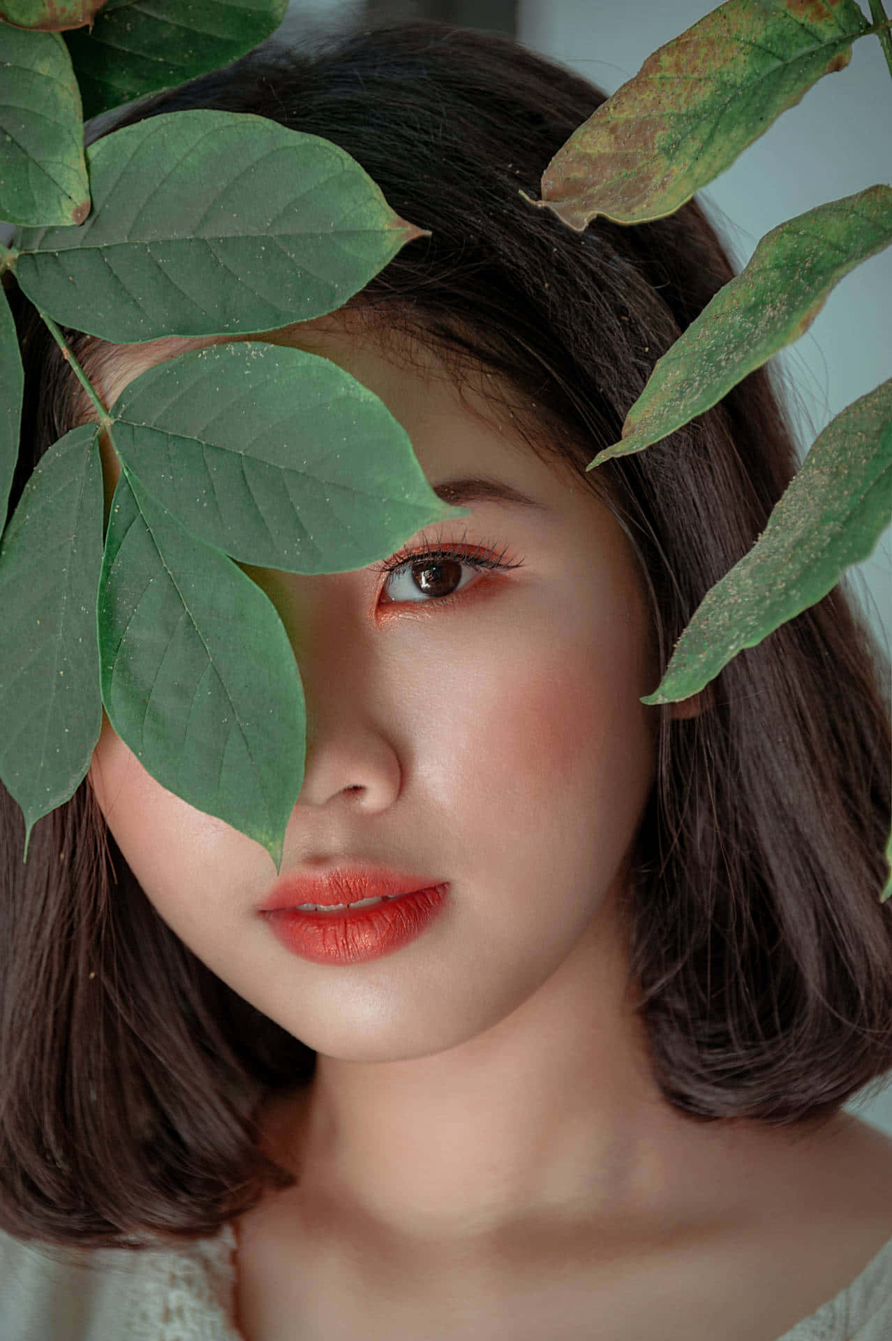  壁紙 — 一位嘴唇鮮紅、露出一隻眼睛的女性，在柔和自然光肖像中透過茂密綠葉窺視。
