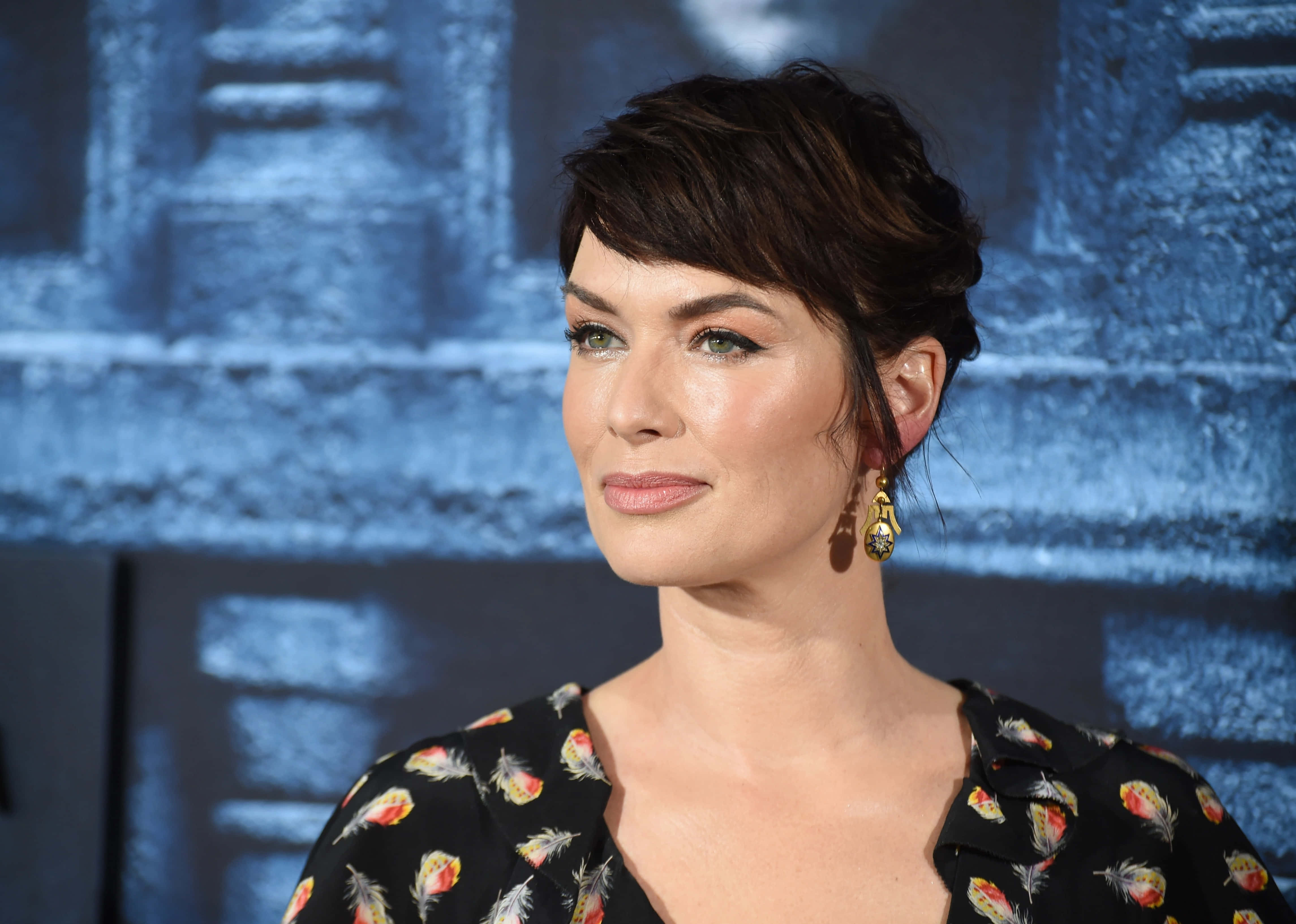 Lena Headey 桌布 壁紙 — 一位短黑髮、身穿羽毛圖案襯衫的女性，在帶有紋理的藍色背景前若有所思地凝視。