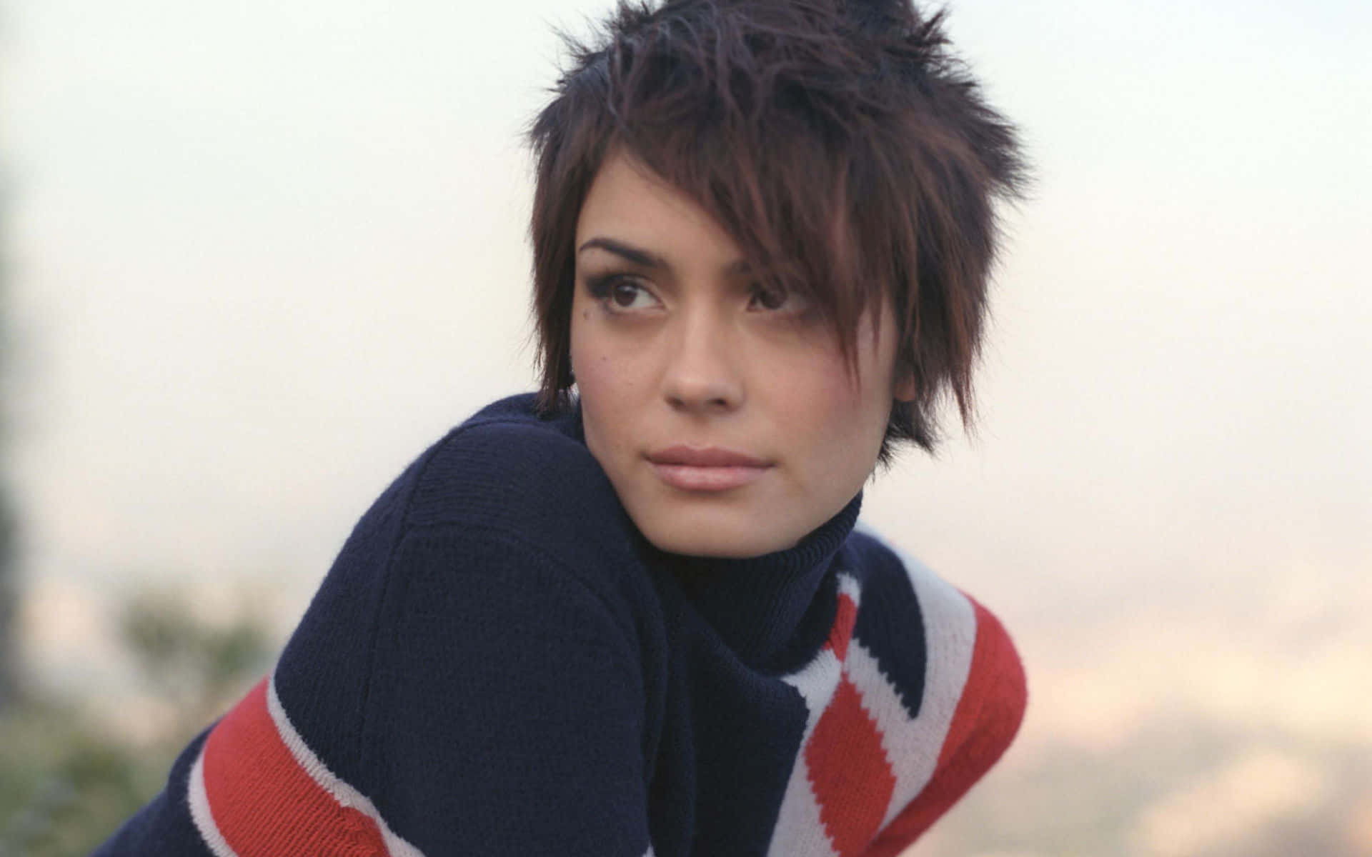 Shannyn Sossamon 桌布 壁紙 — 一位短髮尖刺的女性，穿著帶有紅色和白色幾何條紋的海軍藍毛衣，若有所思地凝視著。