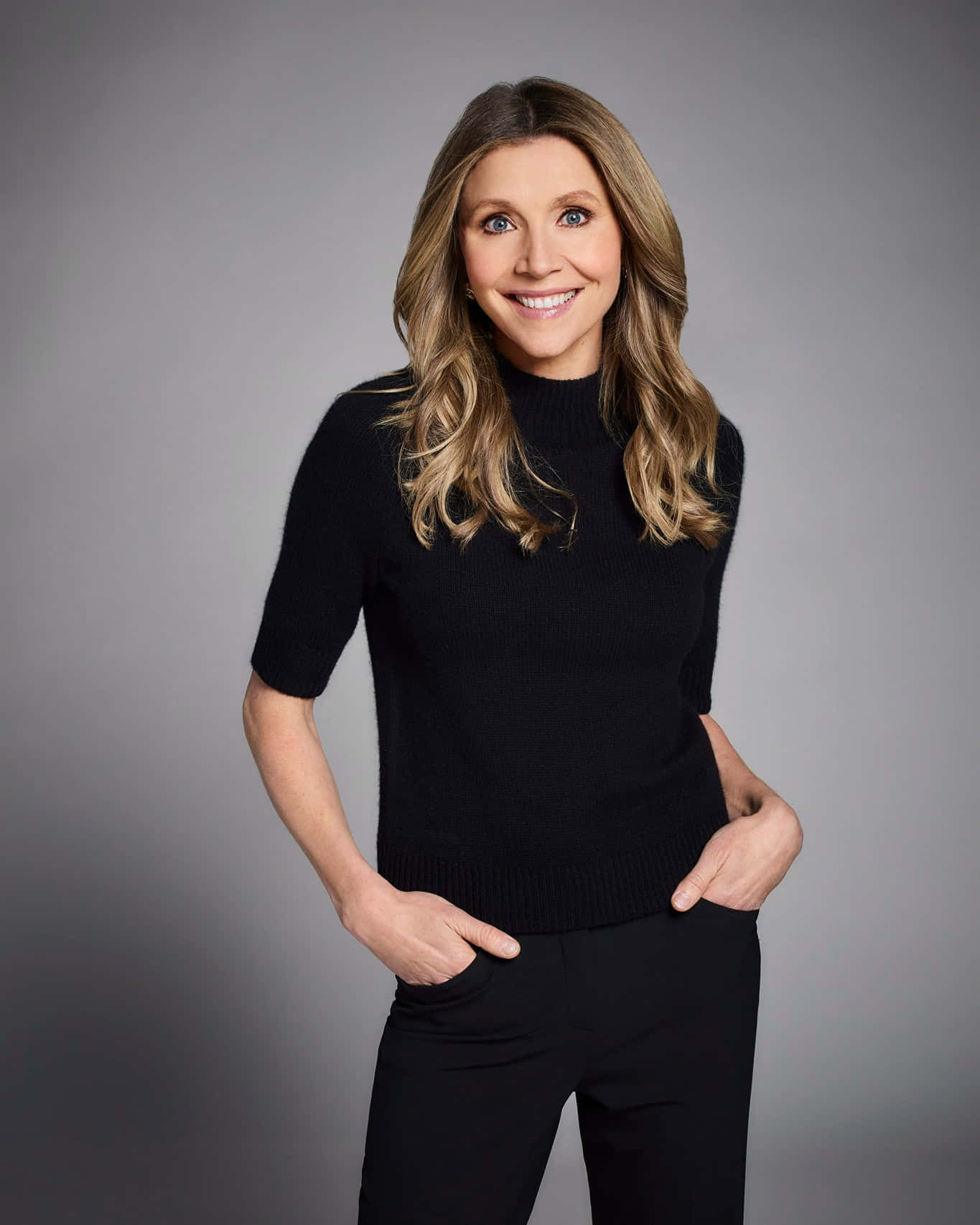 Sarah Chalke 桌布 壁紙 — 一位擁有波浪金髮的女性，穿著黑色毛衣和長褲，在灰色工作室背景前溫暖地微笑。