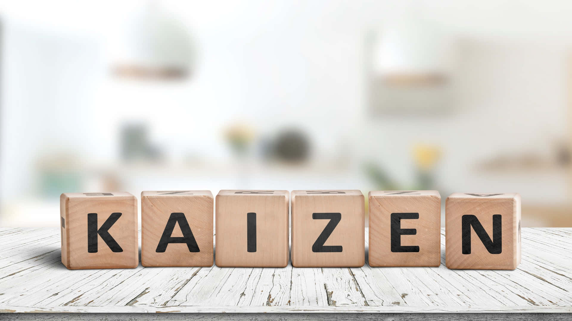  壁紙 — 拼出 KAIZEN 的木塊放在一張風化的白色木桌上，背景是明亮模糊的廚房。
