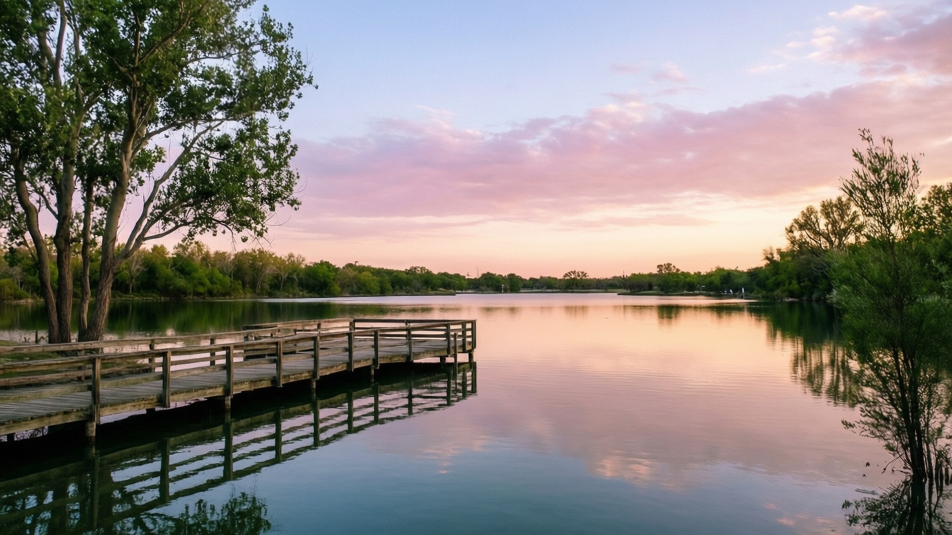 Overholser Lake Sunset 壁紙 — 一個木製碼頭延伸到平靜的湖面，反射著粉紅色和橙色的日落雲彩，周圍環繞著綠樹。