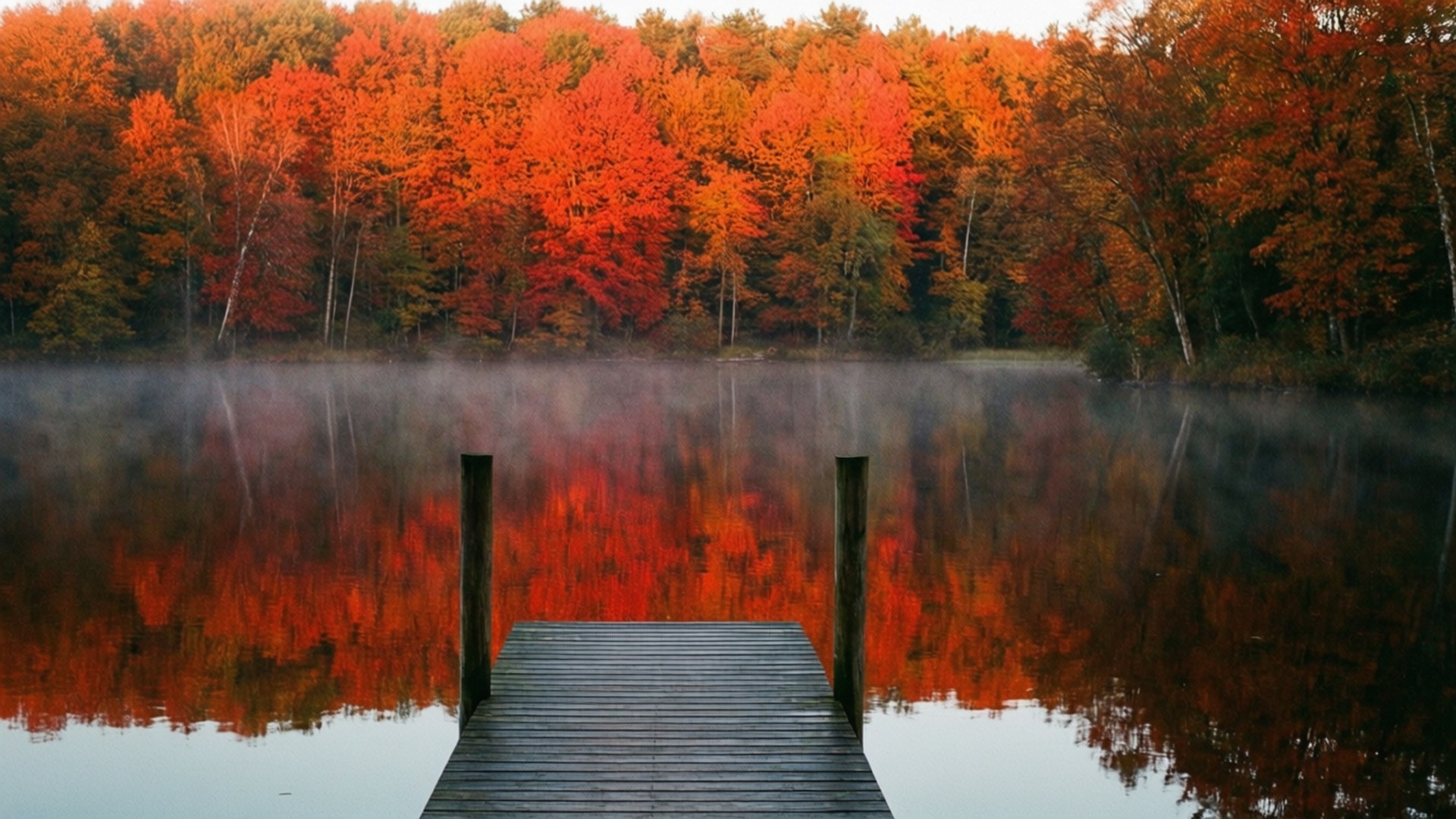 Autumn Lake Reflection 壁紙 — 一個木製碼頭延伸到霧氣瀰漫的湖中，映照著岸邊火紅的秋樹。