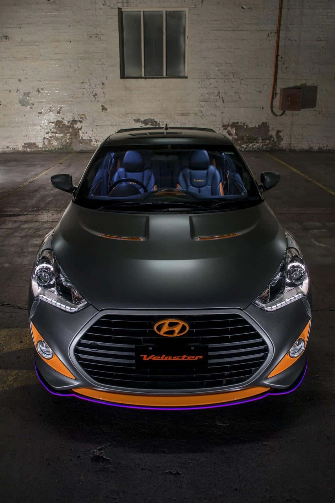 Hyundai Veloster 桌布 壁紙 — 一輛霧面黑色現代Veloster帶有橙色和紫色點綴，在昏暗的工業車庫中正面朝前。