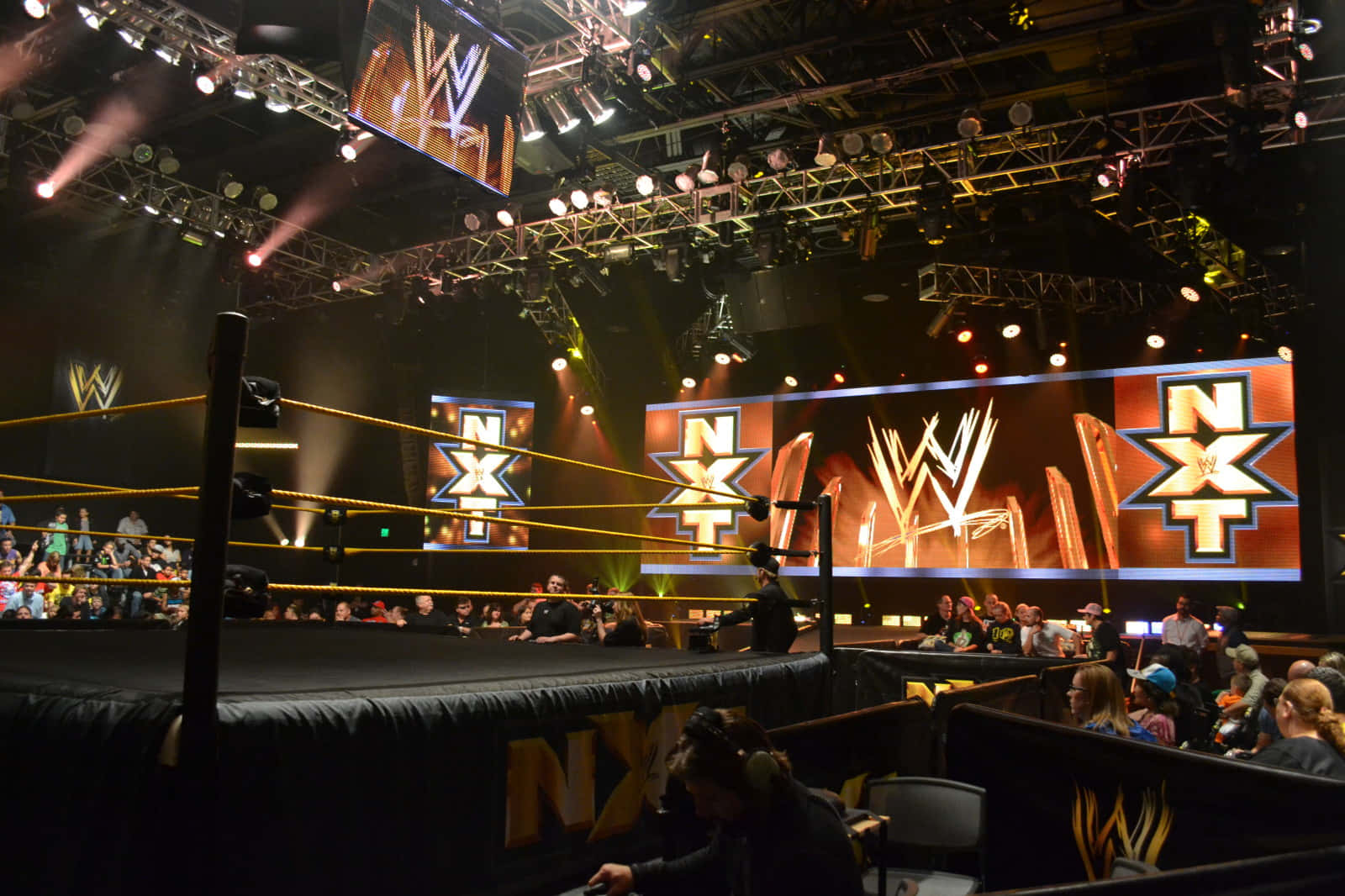 Wrestling Ring 桌布 壁紙 — WWE NXT 摔角擂台在明亮的舞台燈光下，前景有粉絲，上方有大型品牌螢幕。