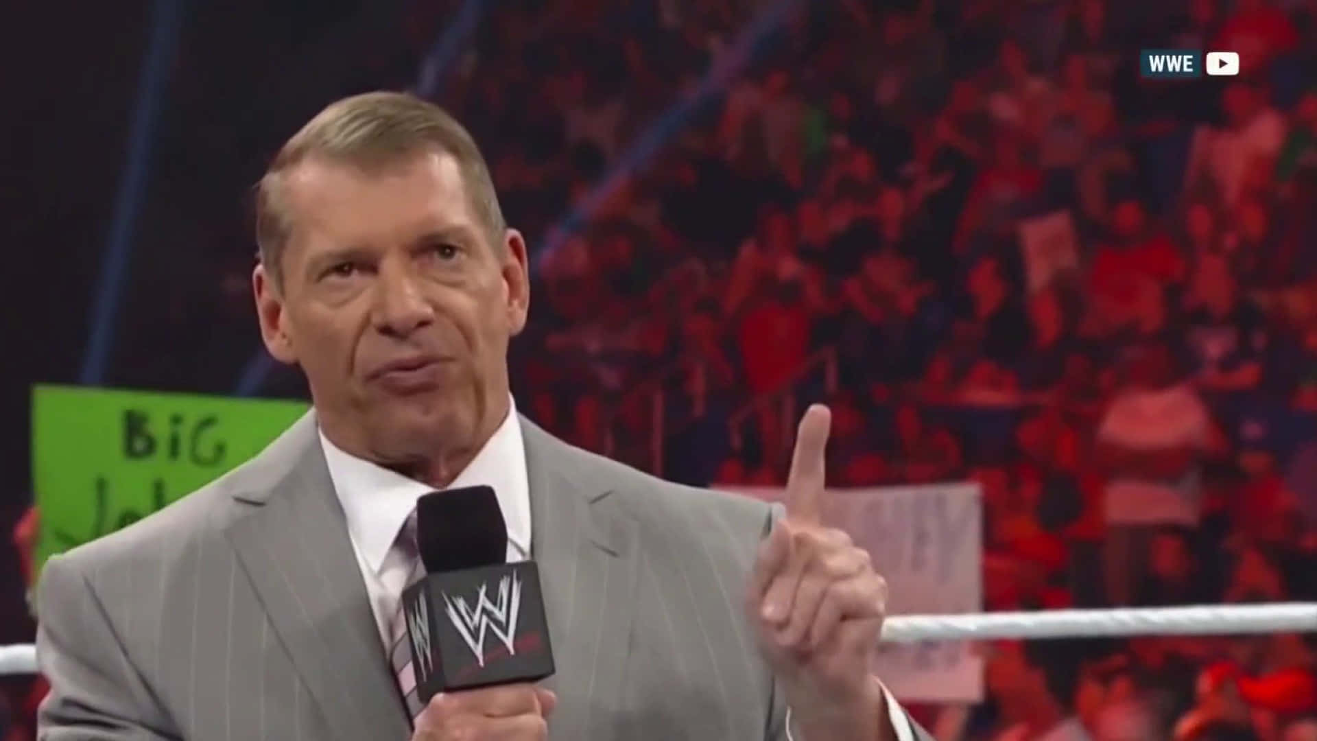 摔角 桌布 壁紙 — WWE 評論員 Vince McMahon 身穿灰色西裝，手持麥克風，在摔角擂台上向上指，身後有模糊的人群。