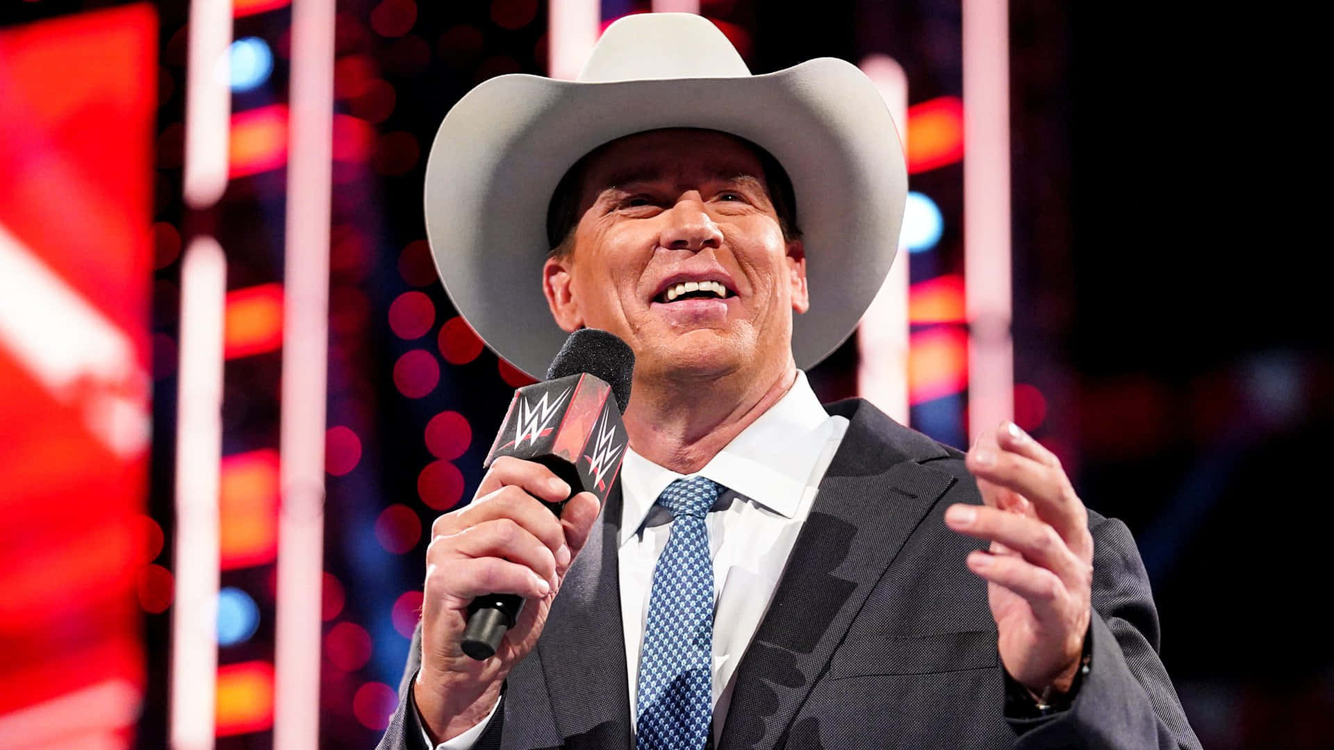 WWE 桌布 壁紙 — WWE 評論員戴著白色牛仔帽和西裝，在鮮紅和藍色燈光舞台背景下拿著麥克風大笑。