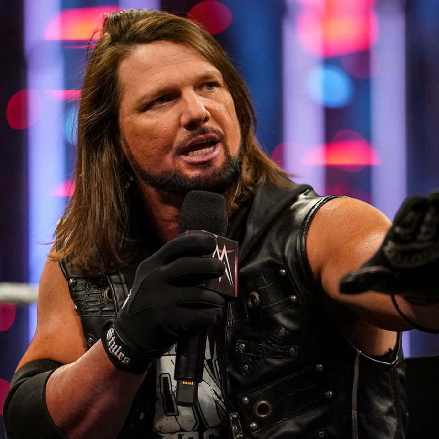 WWE 桌布 壁紙 — WWE摔角手AJ斯泰爾斯穿著黑色皮背心和手套，在色彩繽紛模糊的舞台背景下，熱情地對著麥克風說話。