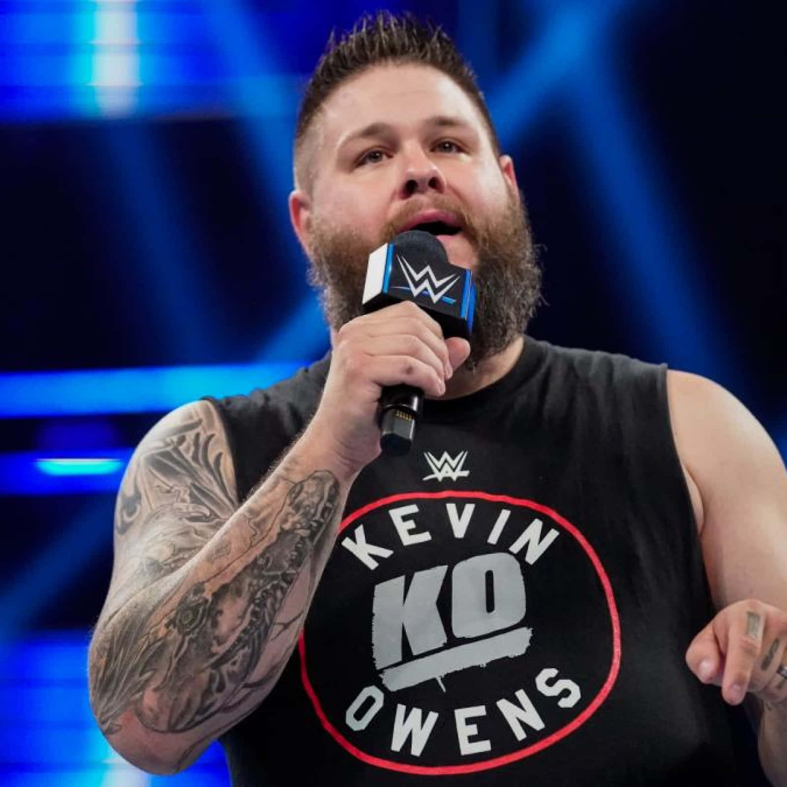 桌布 壁紙 — WWE摔角手Kevin Owens留鬍鬚及紋身，身穿印有他名字的黑色無袖衫，在藍色燈光舞台背景下對著麥克風說話。