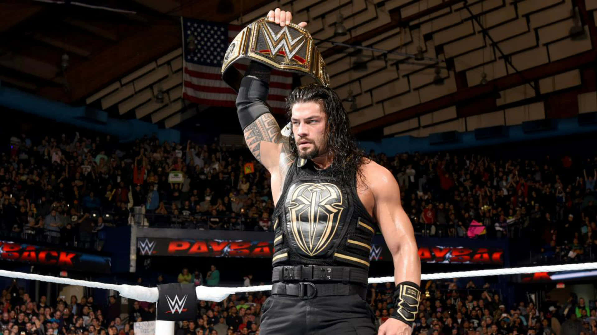 WWE 桌布 壁紙 — WWE摔角手Roman Reigns在明亮的燈光下，於座無虛席的場館中高舉冠軍腰帶，展現勝利姿態。