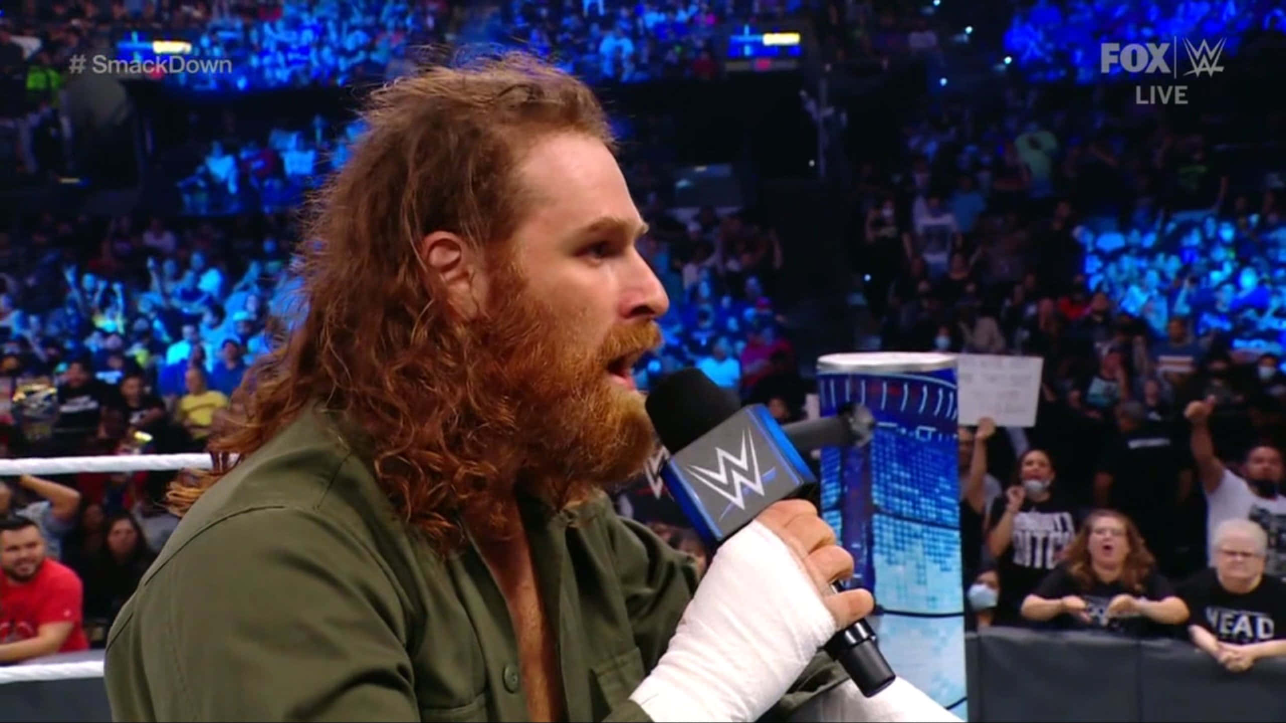 sami zayn 桌布 壁紙 — WWE摔角手留著長紅髮和鬍鬚，在擠滿觀眾的場館內於直播期間對著麥克風說話。