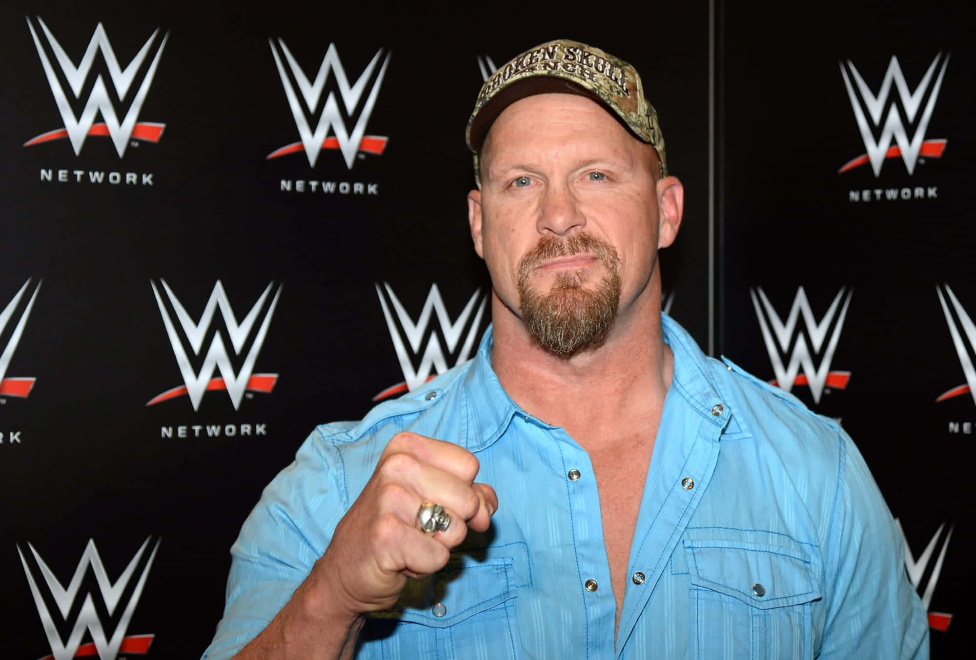 WWE 桌布 壁紙 — WWE摔角手留鬍鬚戴帽，身穿藍色襯衫，在品牌背景前自信擺姿勢，展示戒指。